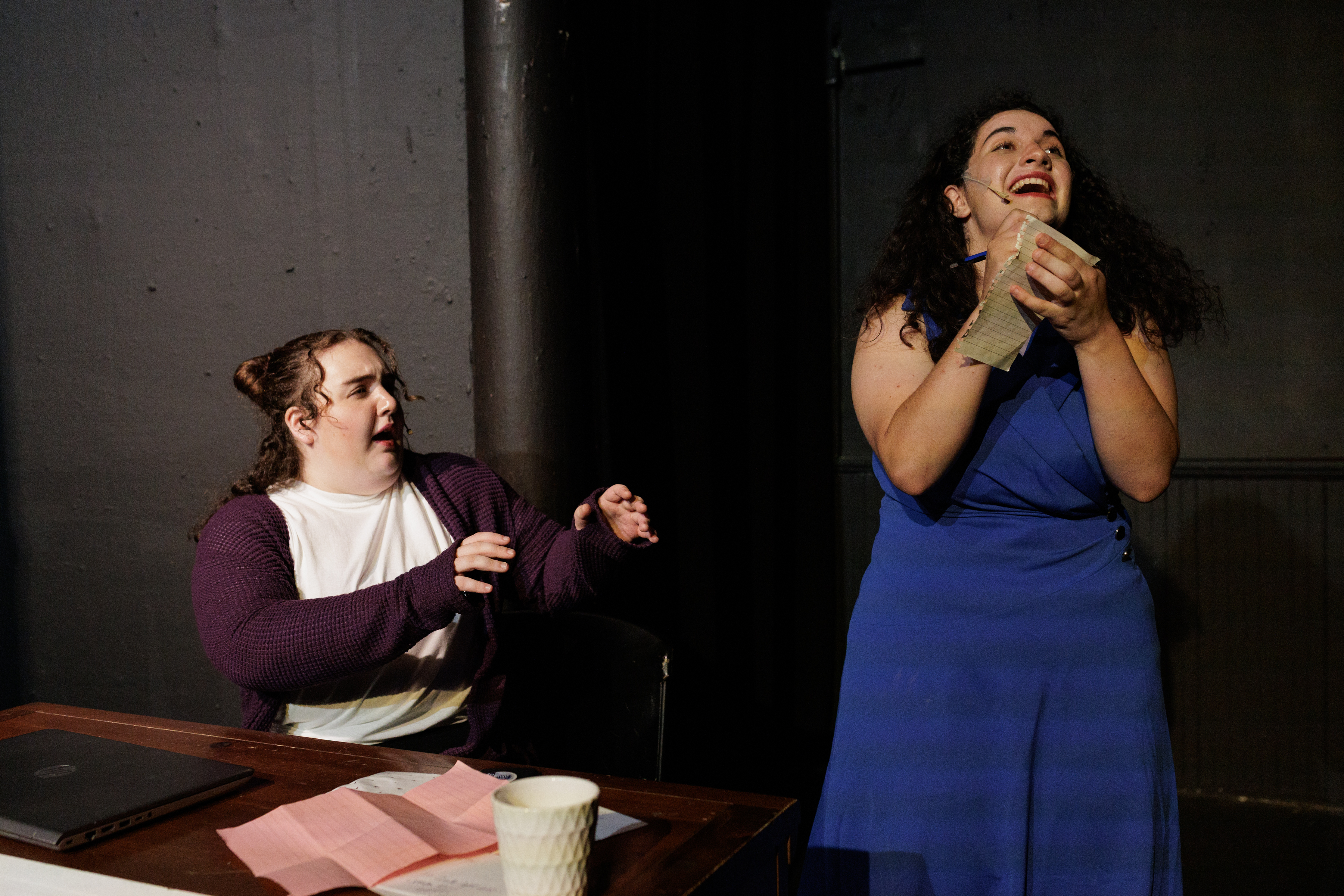 A performer in blue clutches papers and looks upward as a seated performer reaches toward them in Two Part Tragedy.
