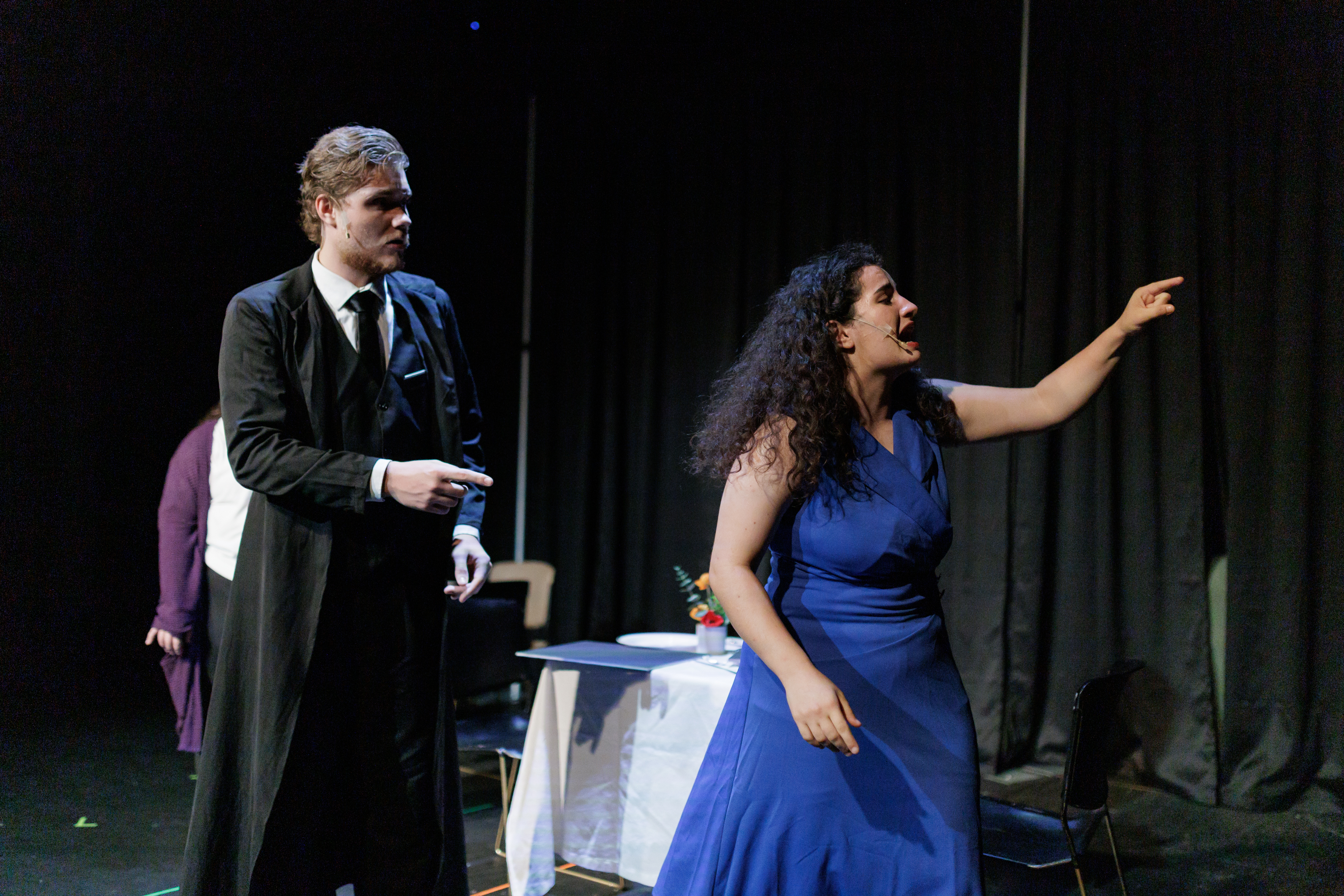 A performer in a blue dress points across the stage beside a performer in a black suit and long coat in Two Part Tragedy.