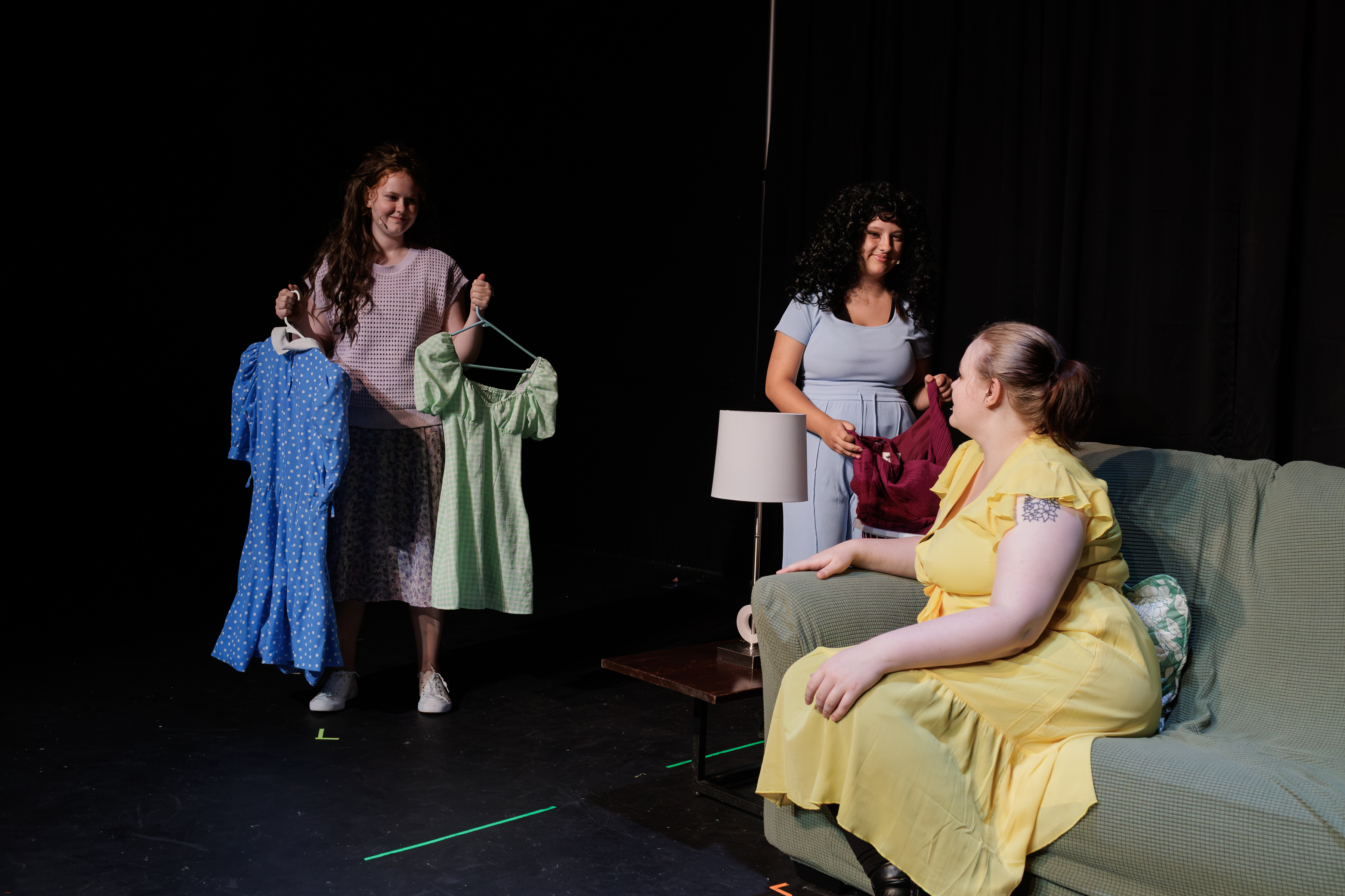 A performer holds blue and green dresses on hangers while two others gather beside a green sofa in Two Part Tragedy.