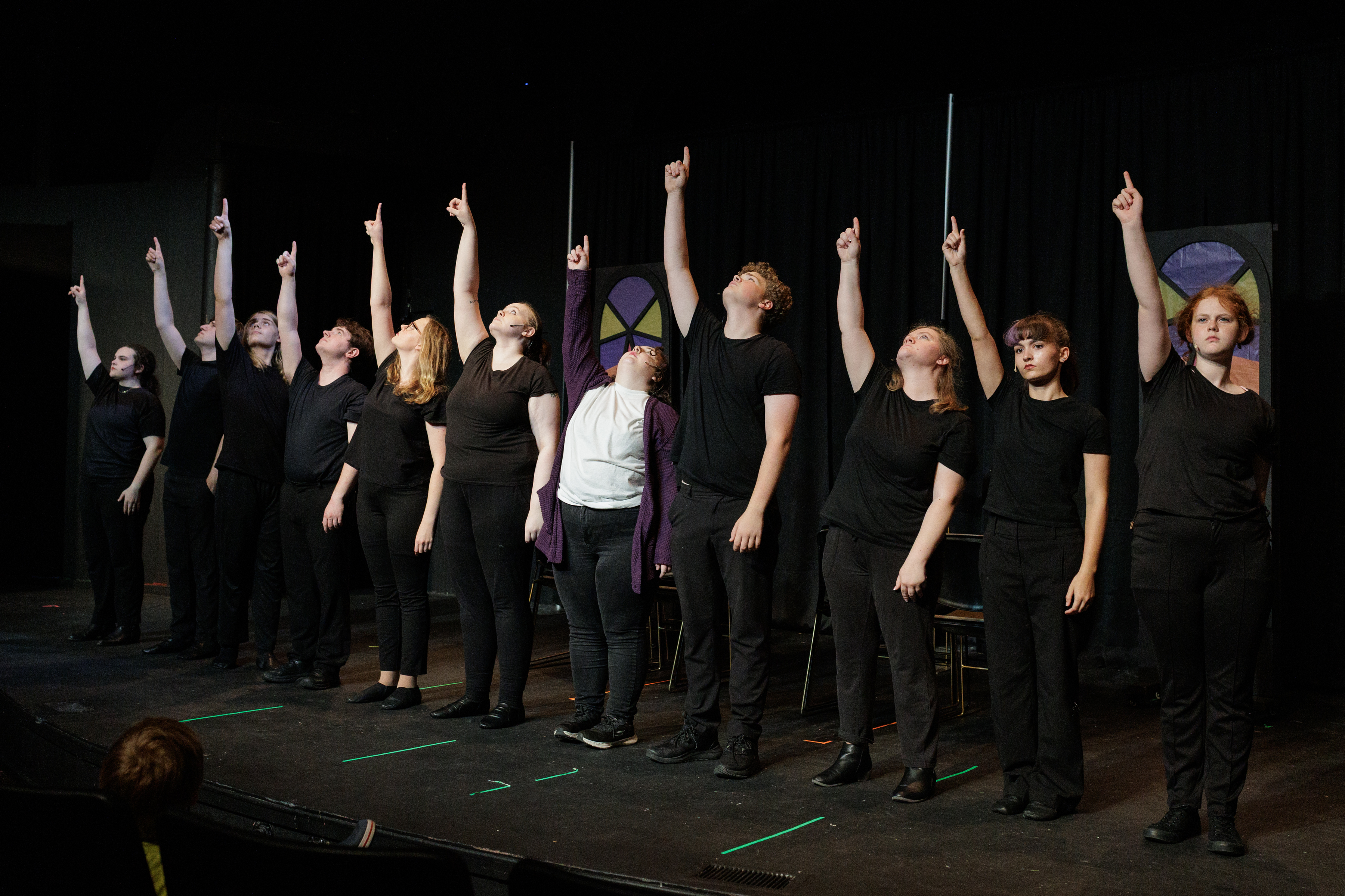 An ensemble stands in a line, looking upward with index fingers raised, in Two Part Tragedy.