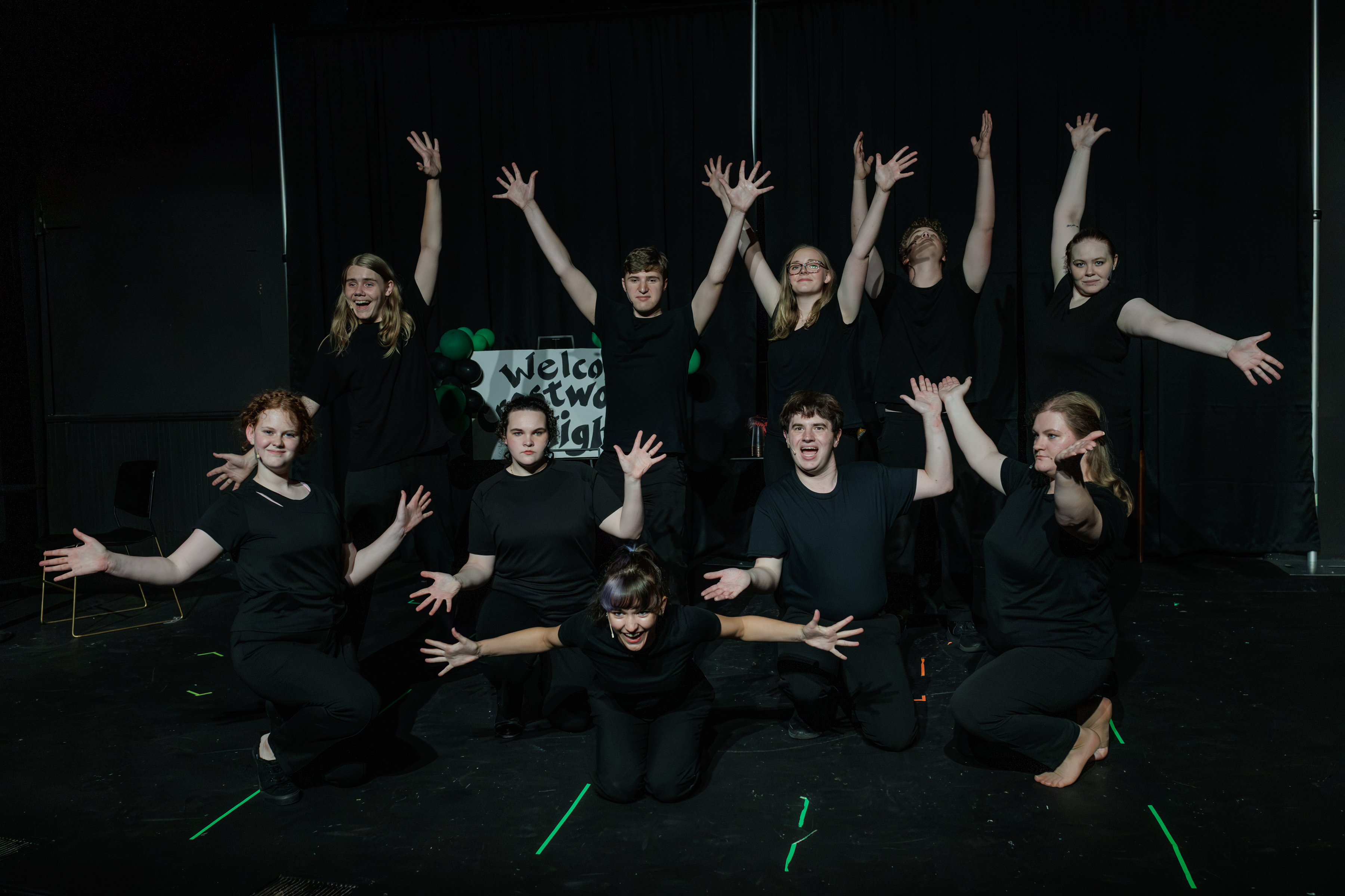 Black-clad performers kneel and stand with arms spread or raised in a group tableau in Two Part Tragedy.