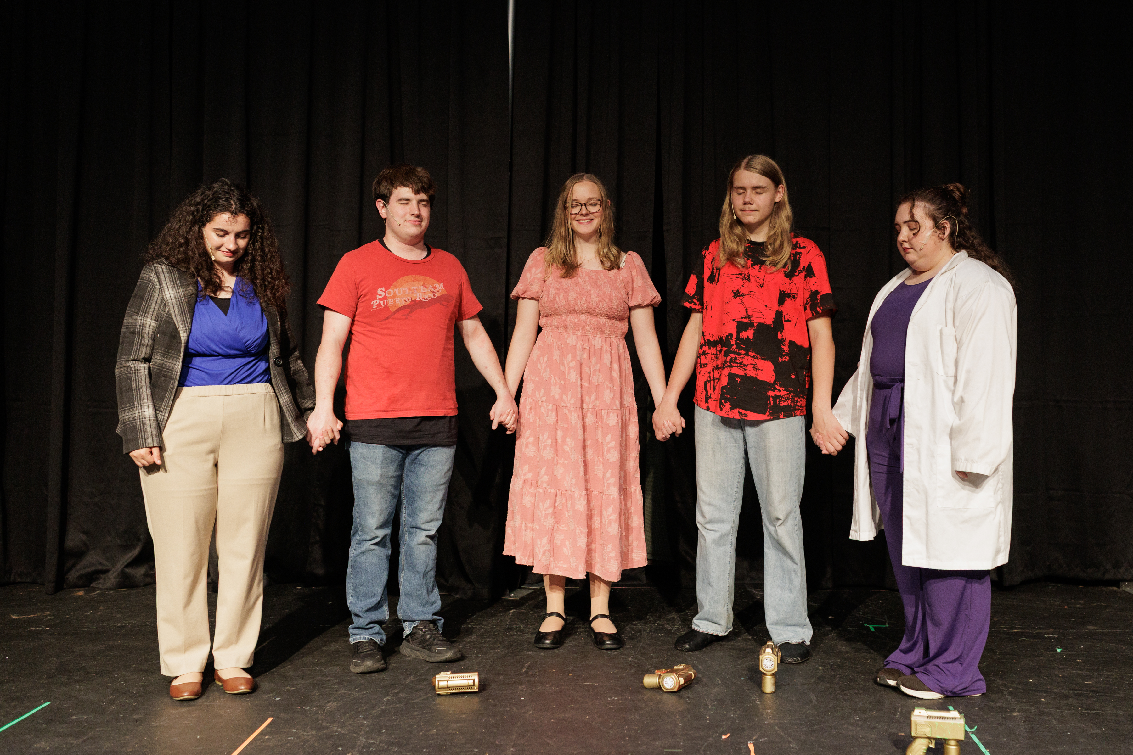Five performers hold hands in a row with small props scattered at their feet in Two Part Tragedy.
