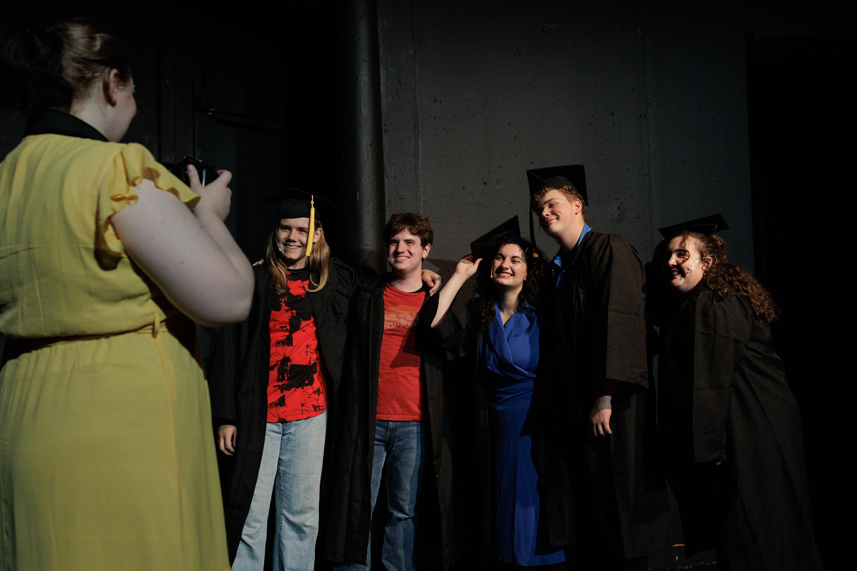Performers in graduation caps and gowns pose while a performer in yellow holds a camera in Two Part Tragedy.