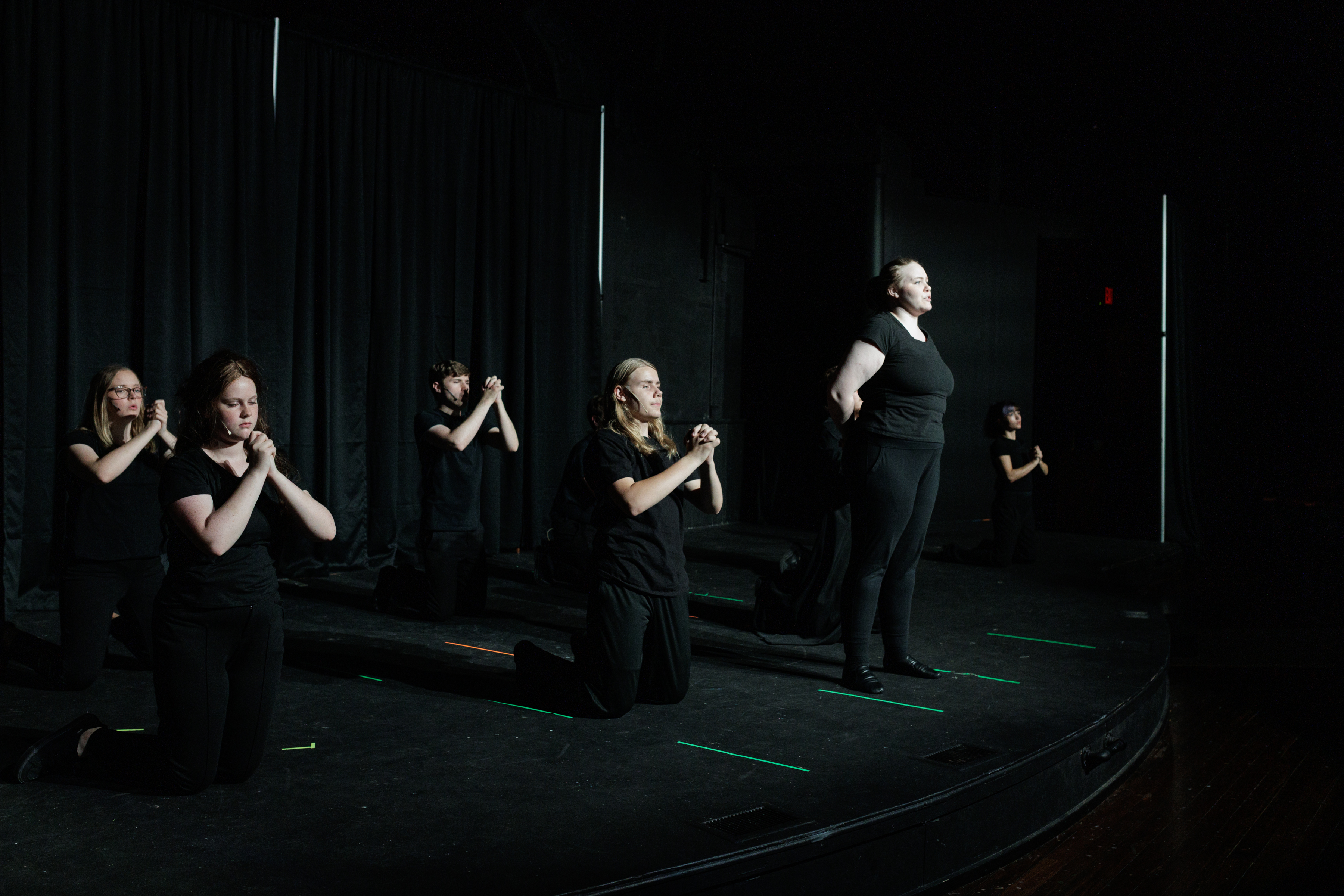 Black-clad ensemble members kneel with clasped hands beside a standing performer in Two Part Tragedy.