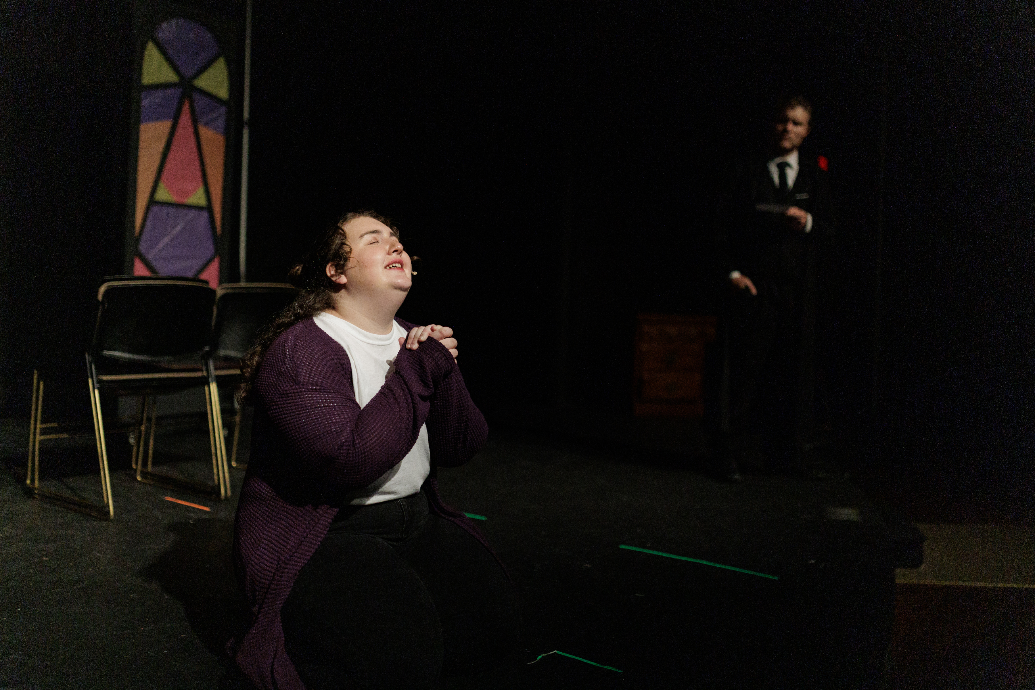 A performer in a purple cardigan kneels with clasped hands and face tilted upward in Two Part Tragedy.