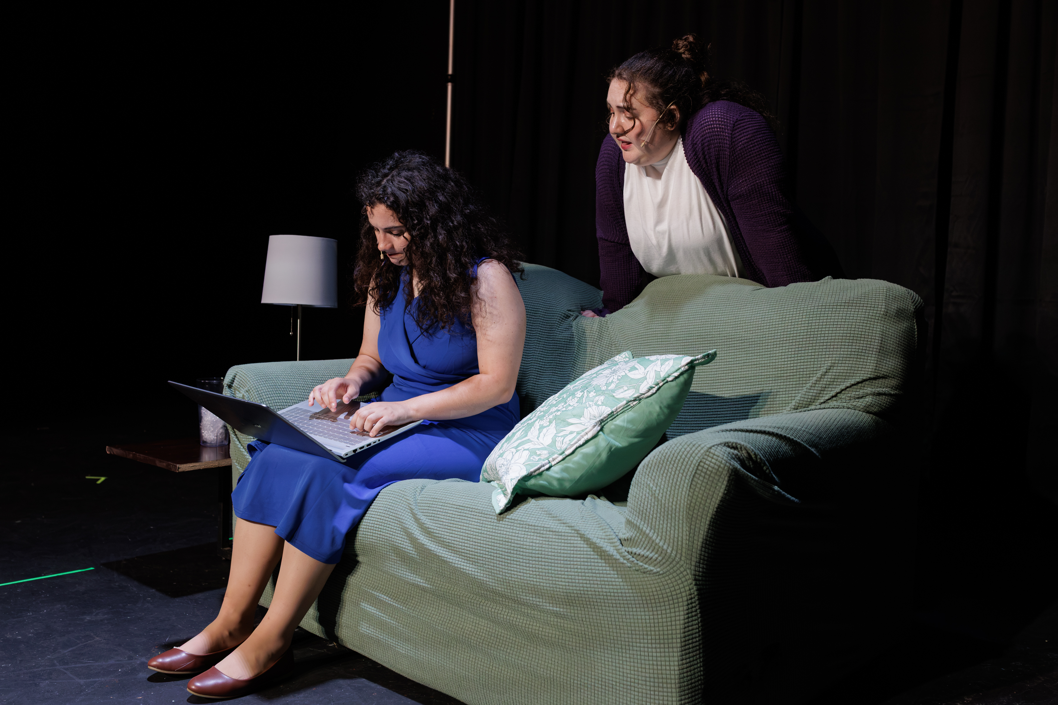 A performer in a blue dress uses a laptop on a green sofa while another leans over the back in Two Part Tragedy.