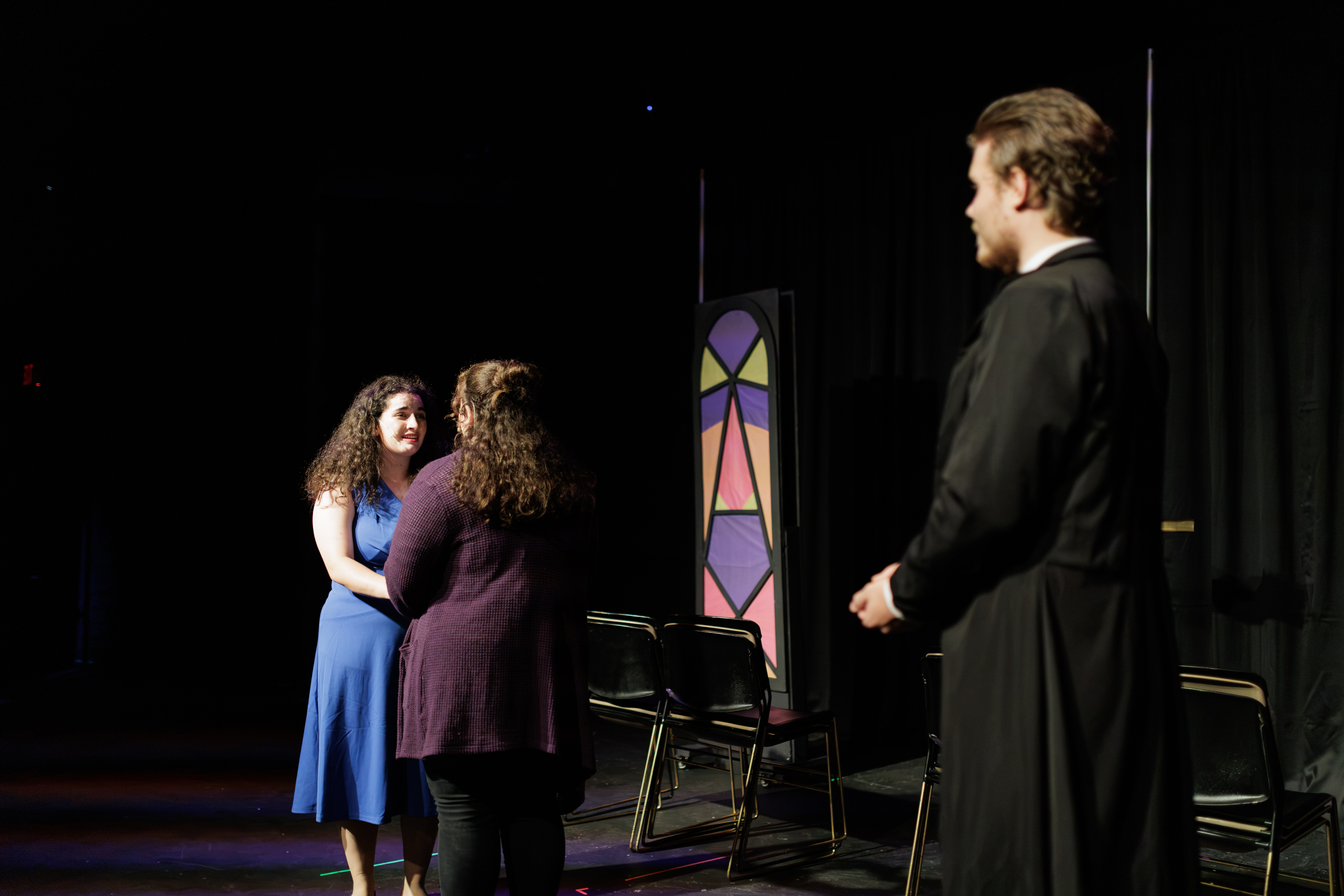 Performers in blue and purple hold hands while a performer in a long black coat stands nearby in Two Part Tragedy.