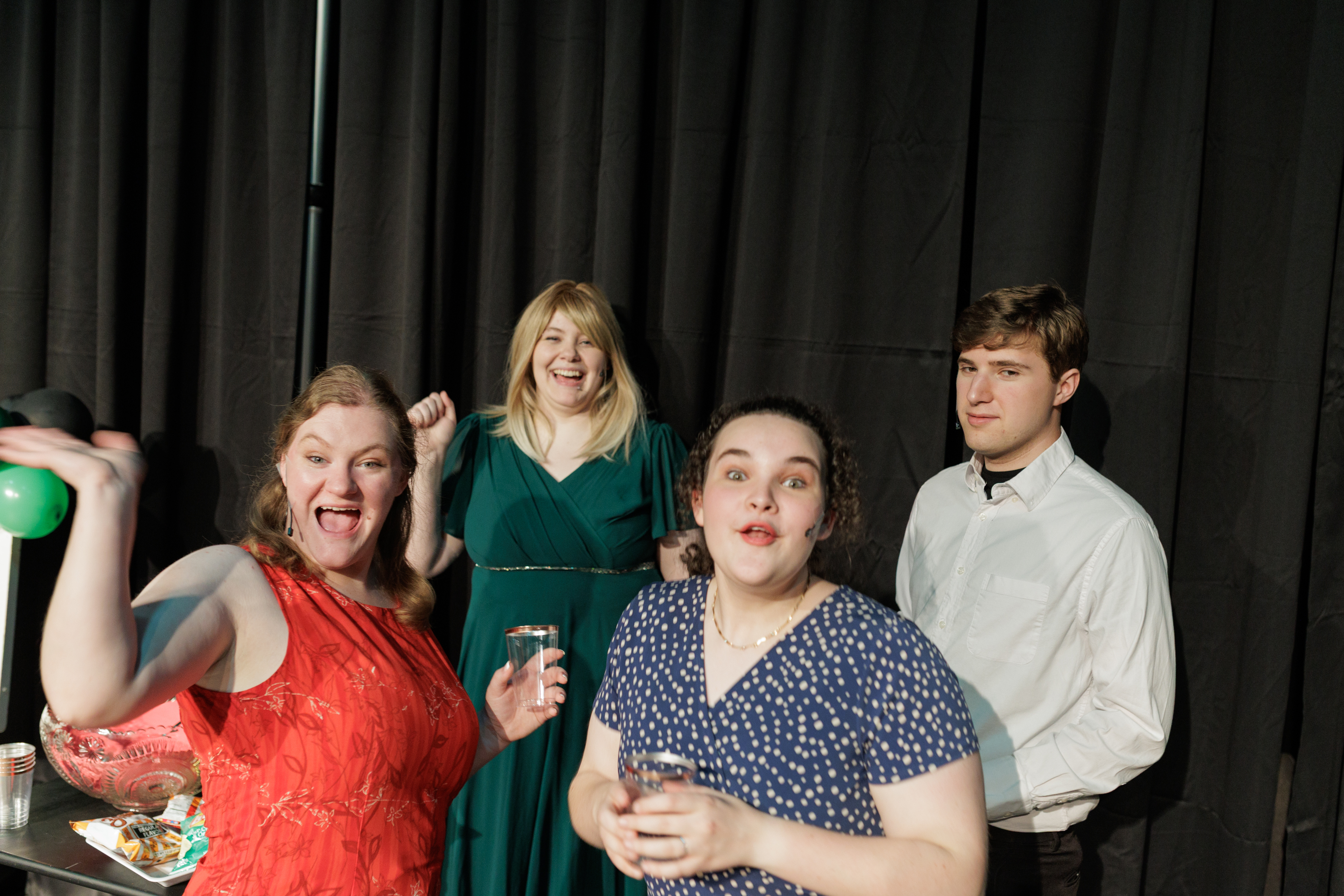 Four performers gather by a refreshment table, with cups and a green balloon, in Two Part Tragedy.