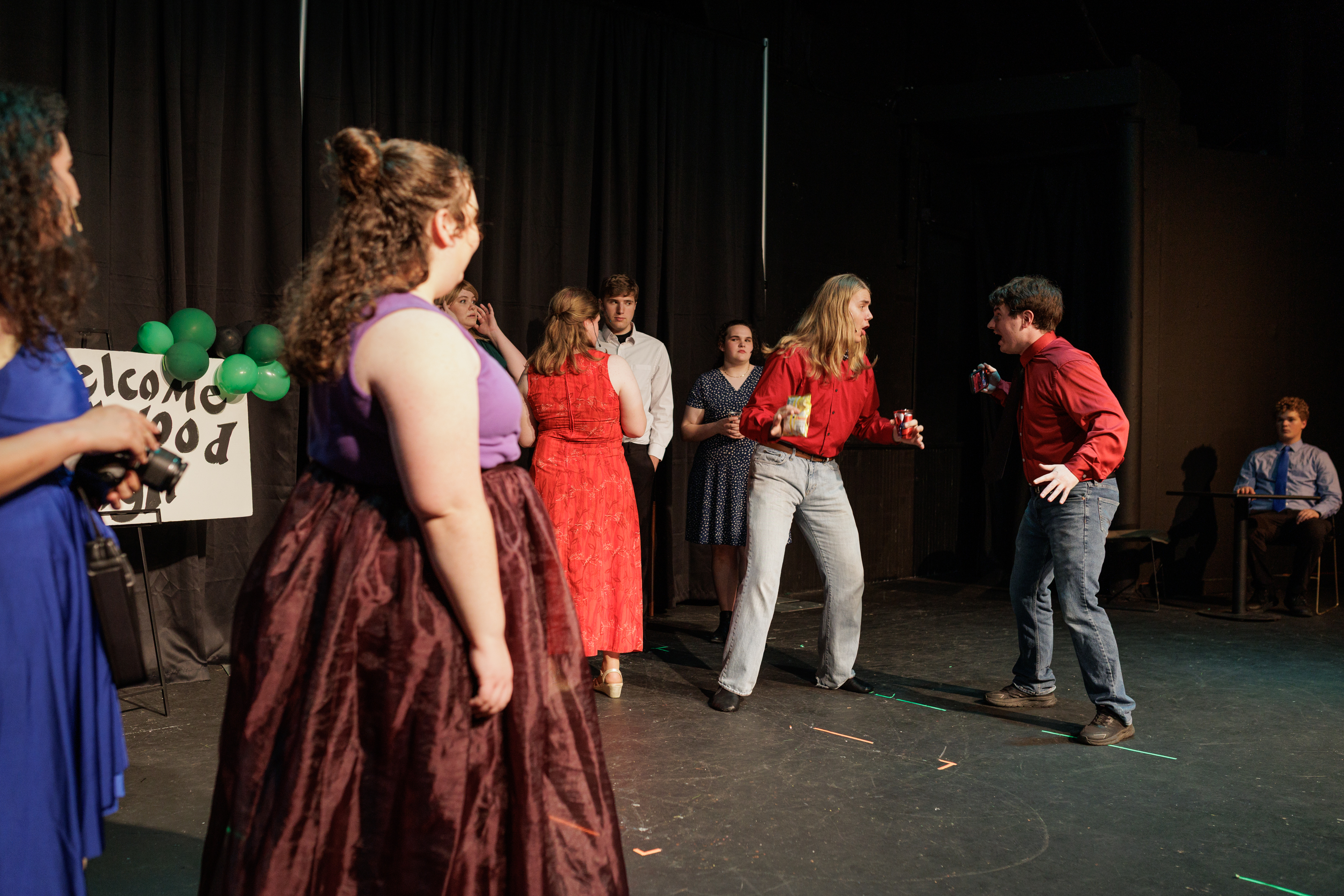 Performers hold cups and dance beside green balloons and a welcome sign in Two Part Tragedy.