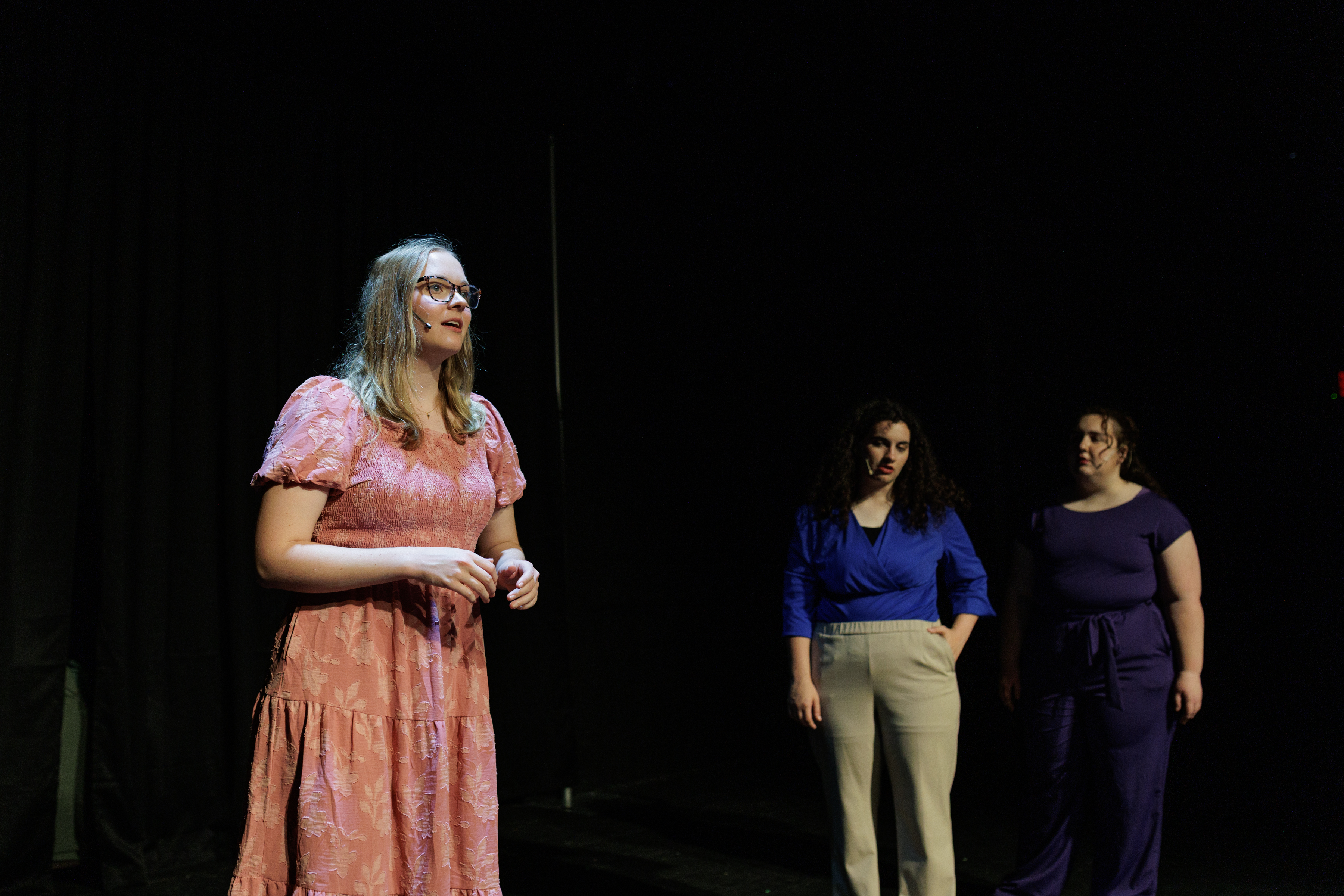 A performer in a pink dress and glasses stands in the foreground with two performers behind in Two Part Tragedy.