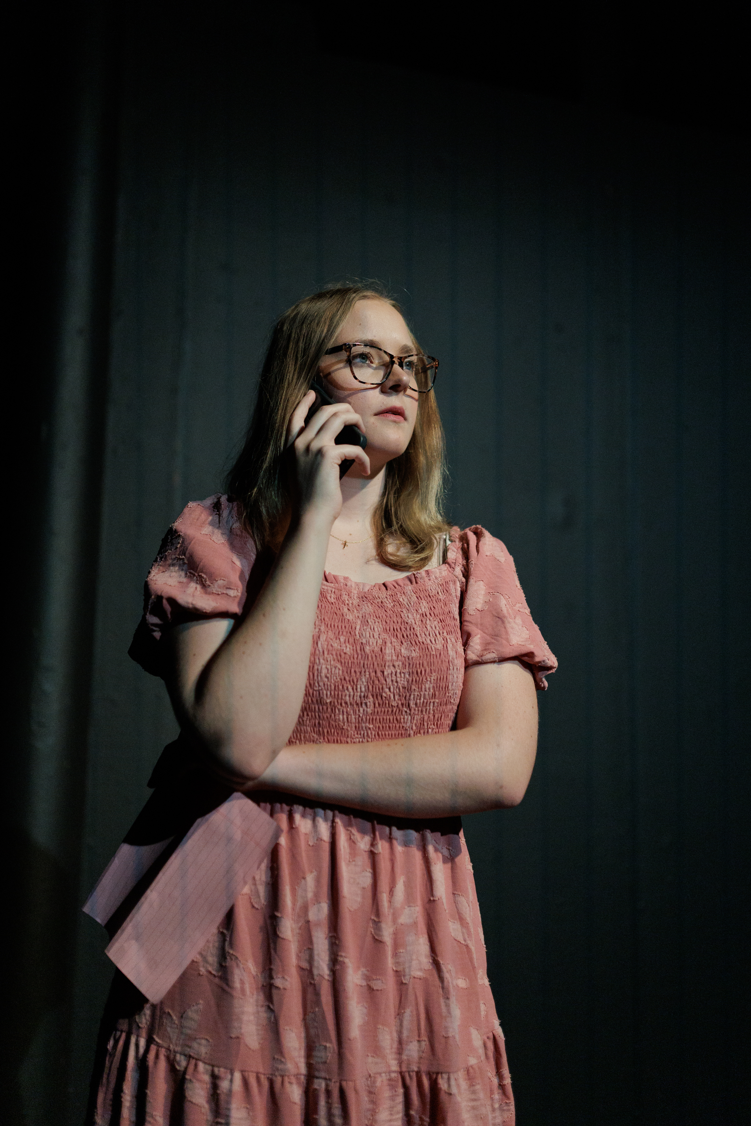 A performer wearing glasses and a pink dress holds a phone to their ear in Two Part Tragedy.