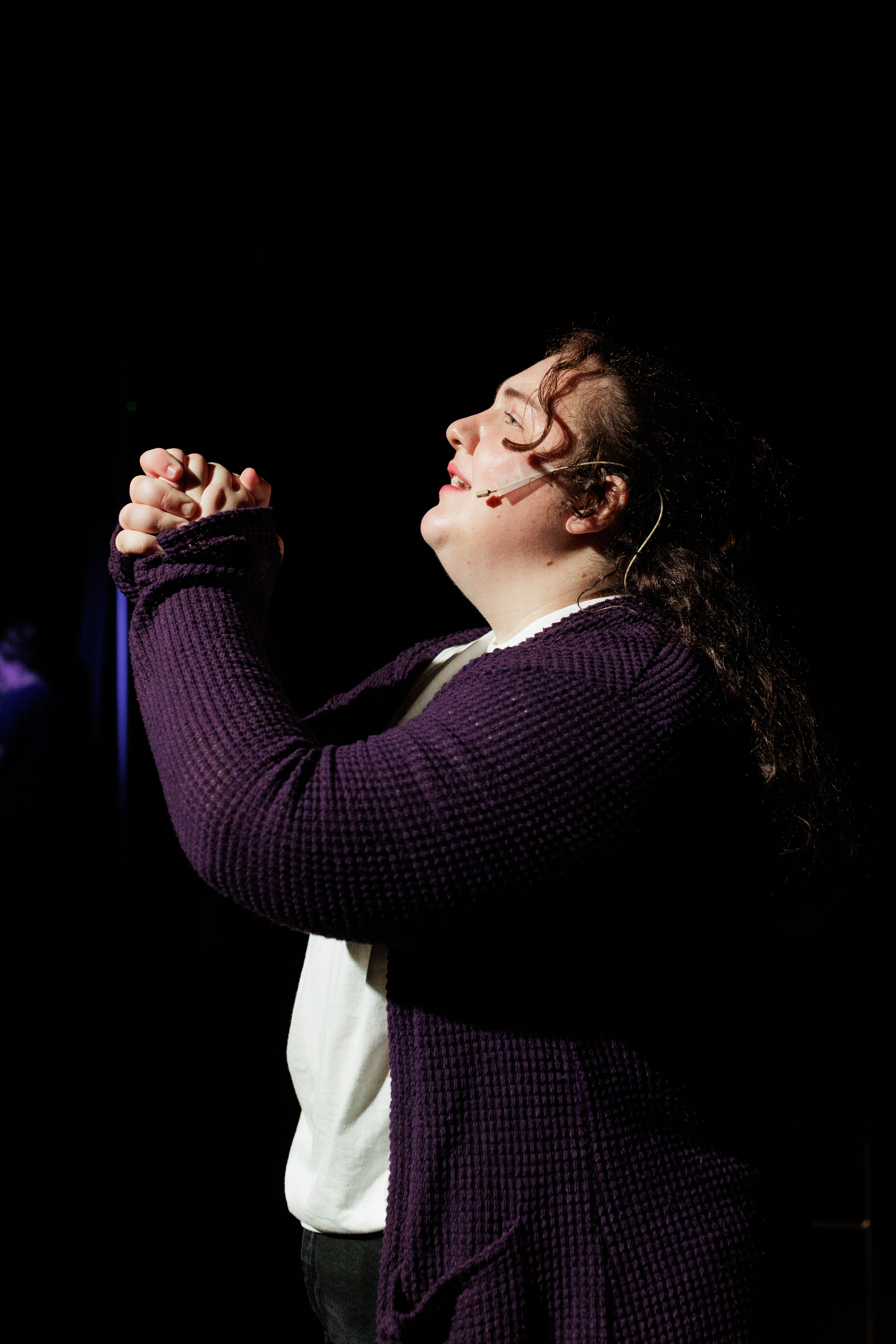 A performer in a purple cardigan raises clasped hands with eyes closed in Two Part Tragedy.
