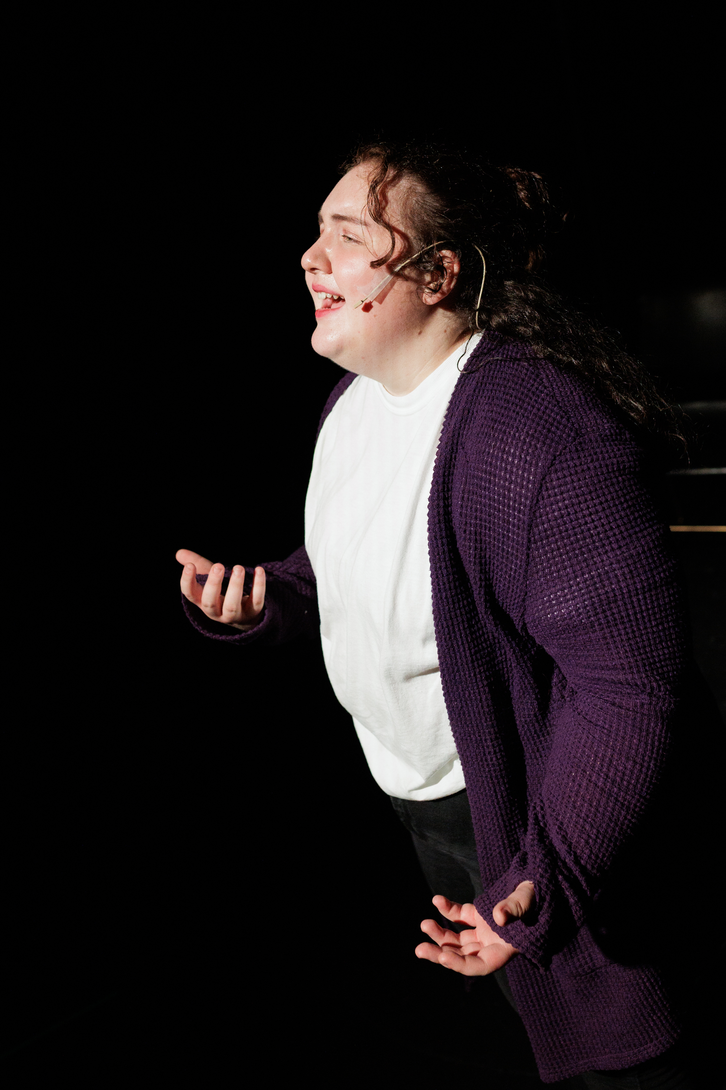 A performer in a purple cardigan speaks with an open hand raised in Two Part Tragedy.
