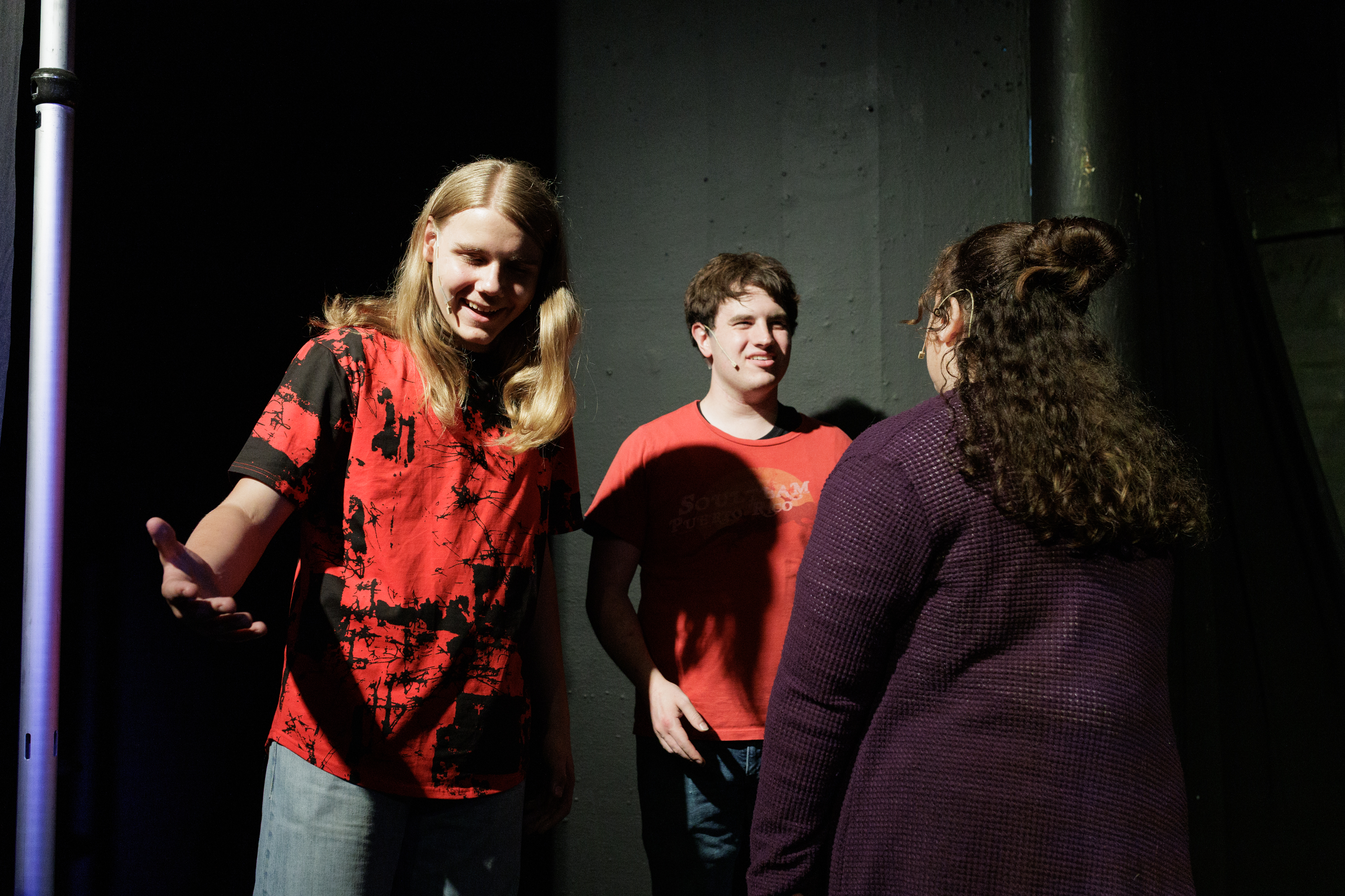 Two red-shirted performers face a performer in a purple cardigan, with one extending an open hand, in Two Part Tragedy.