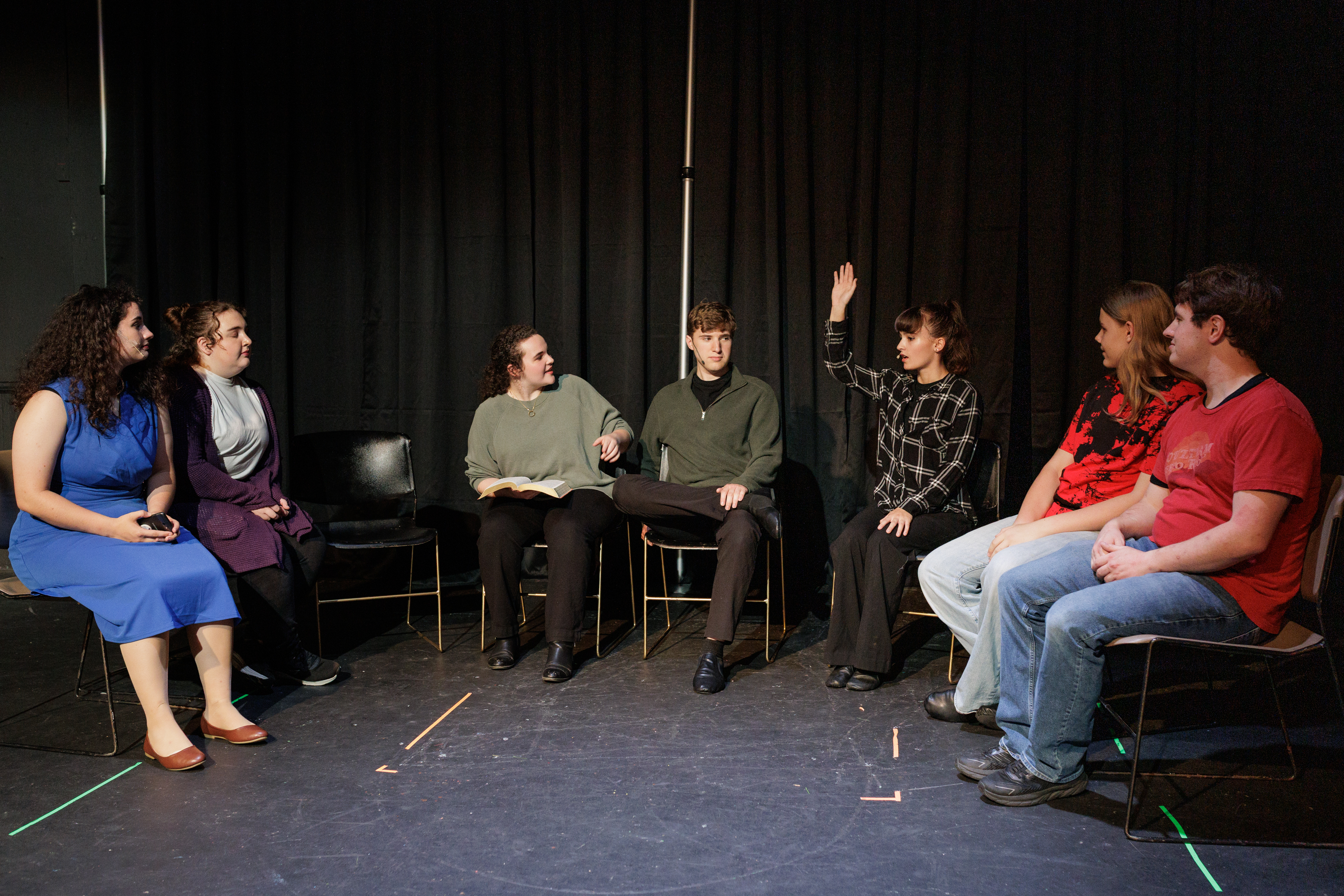 Performers sit in a semicircle as one raises a hand and another holds an open book in Two Part Tragedy.