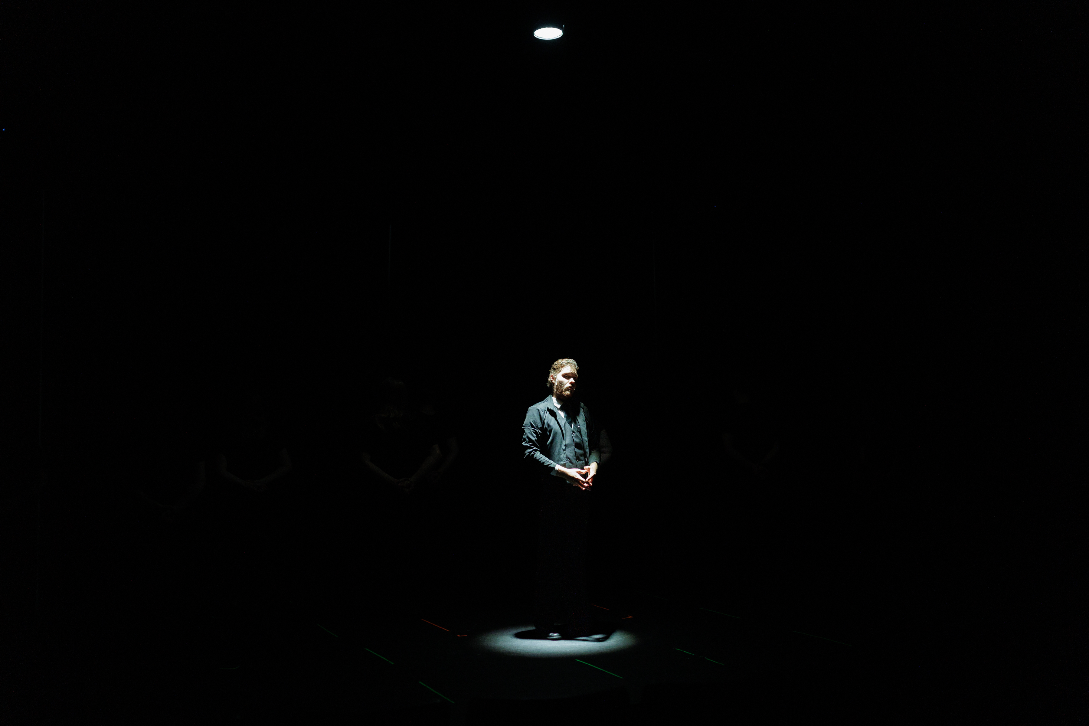 A performer in a dark suit stands in a small spotlight on an otherwise dark stage in Two Part Tragedy.