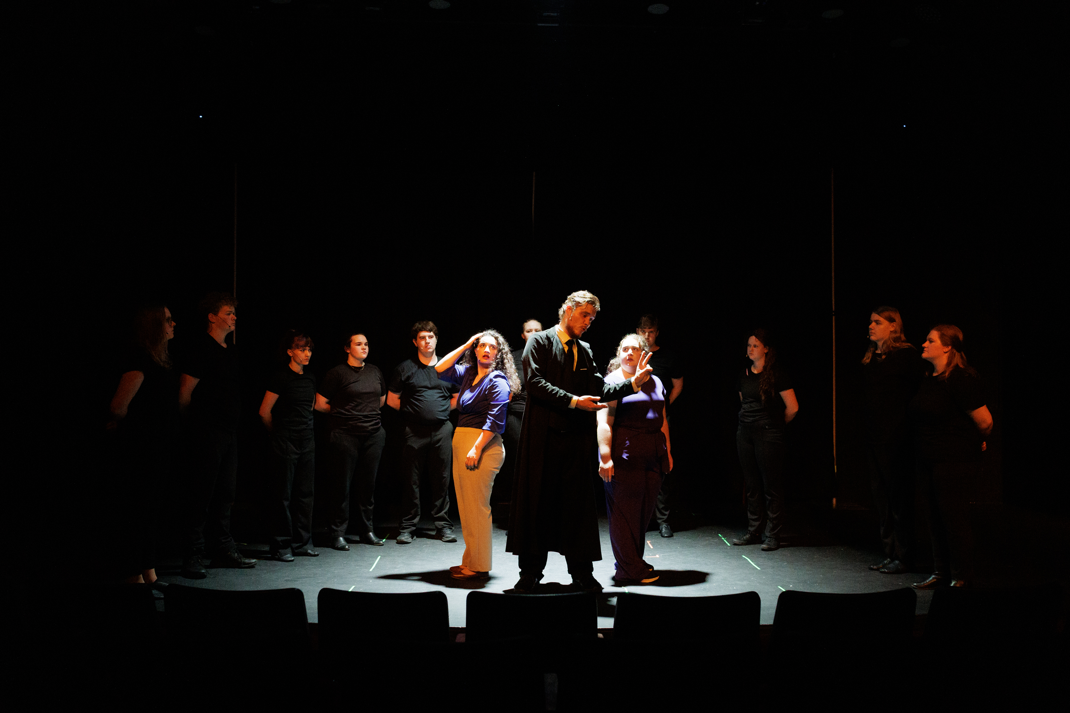 Three performers stand together in a bright pool of light, surrounded by a black-clad ensemble in Two Part Tragedy.