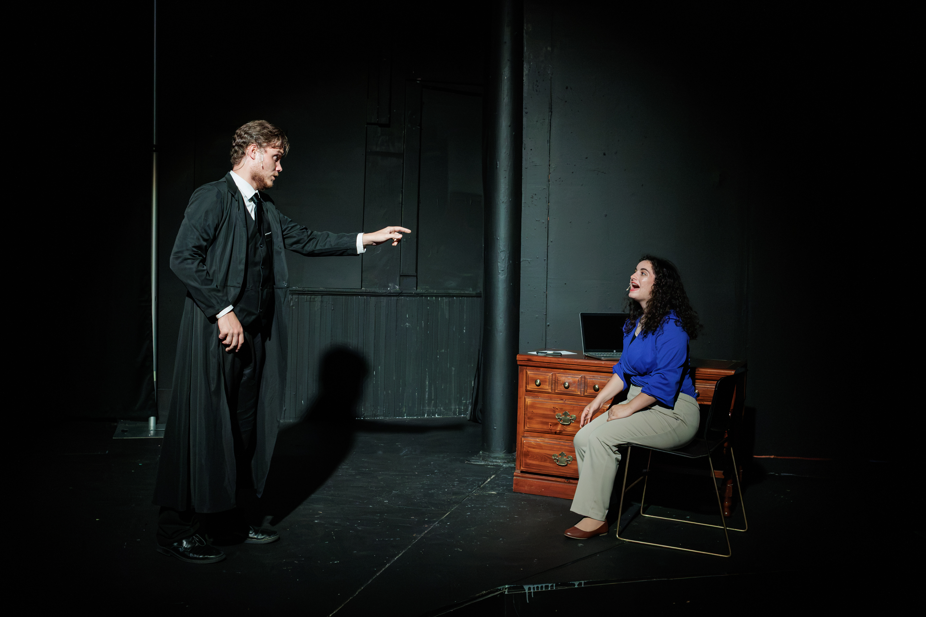 A performer in a long black coat reaches toward a seated performer in a blue blouse beside a desk in Two Part Tragedy.