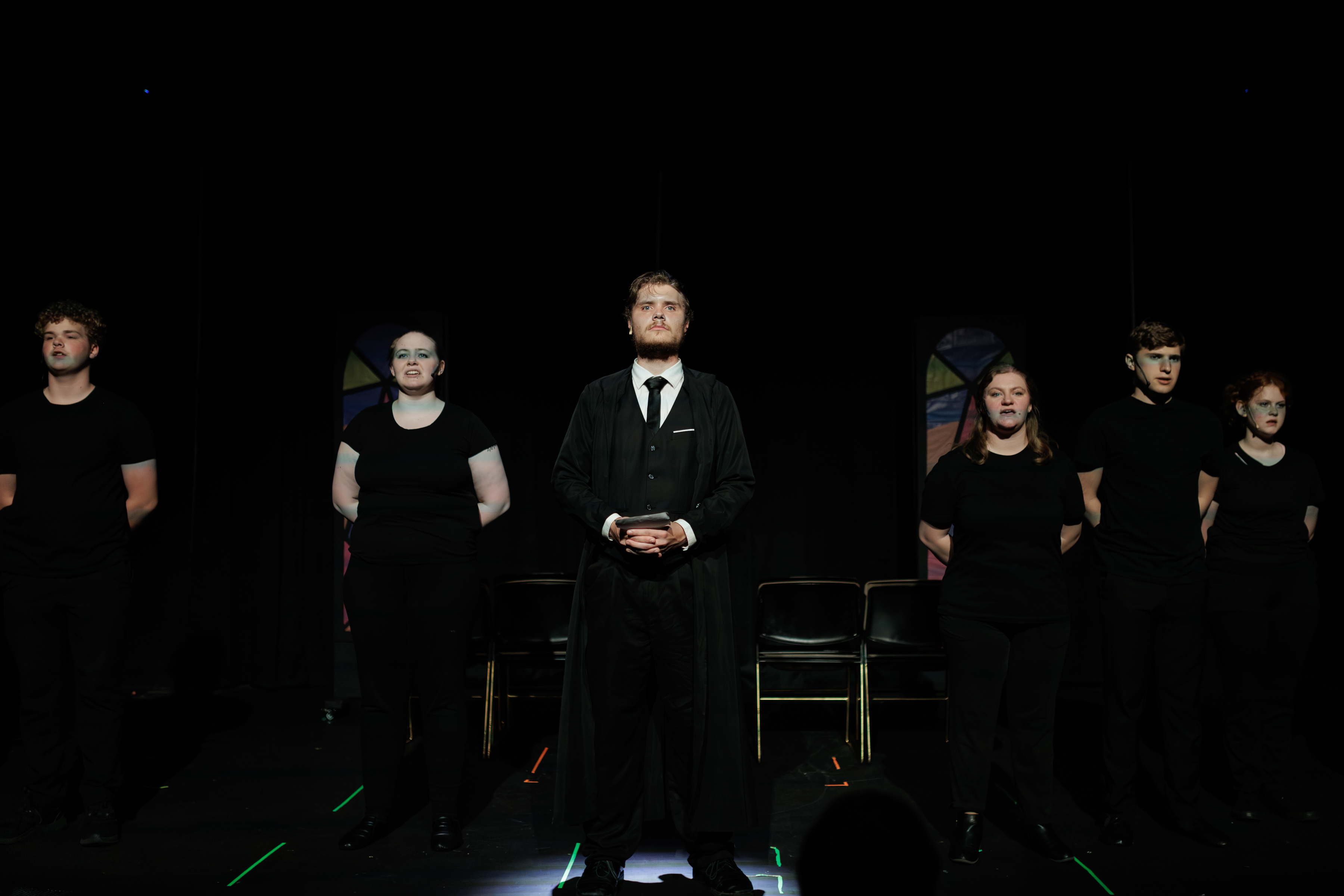 A suited performer stands with hands clasped, surrounded by a black-clad ensemble on a dark stage in Two Part Tragedy.