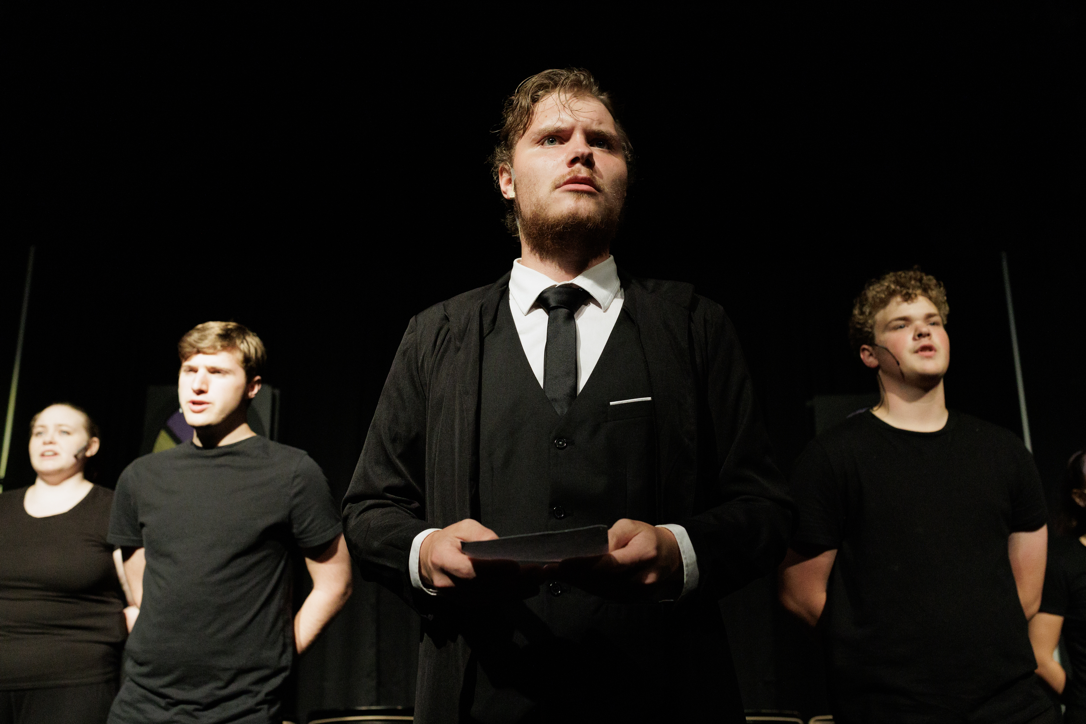 A performer in a black suit and long coat holds a closed book with black-clad performers behind in Two Part Tragedy.