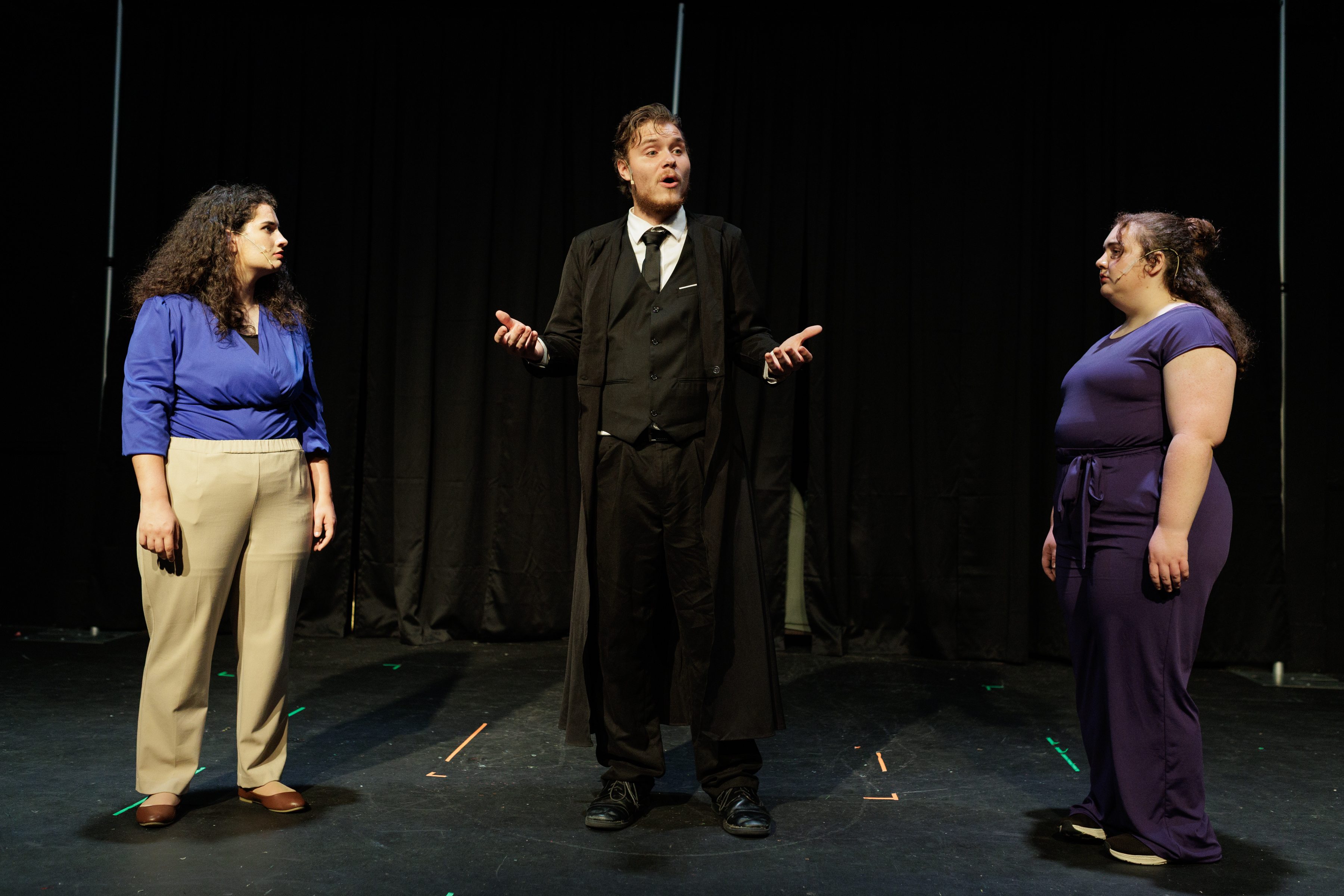A suited performer holds both palms open between performers in a blue blouse and purple outfit in Two Part Tragedy.