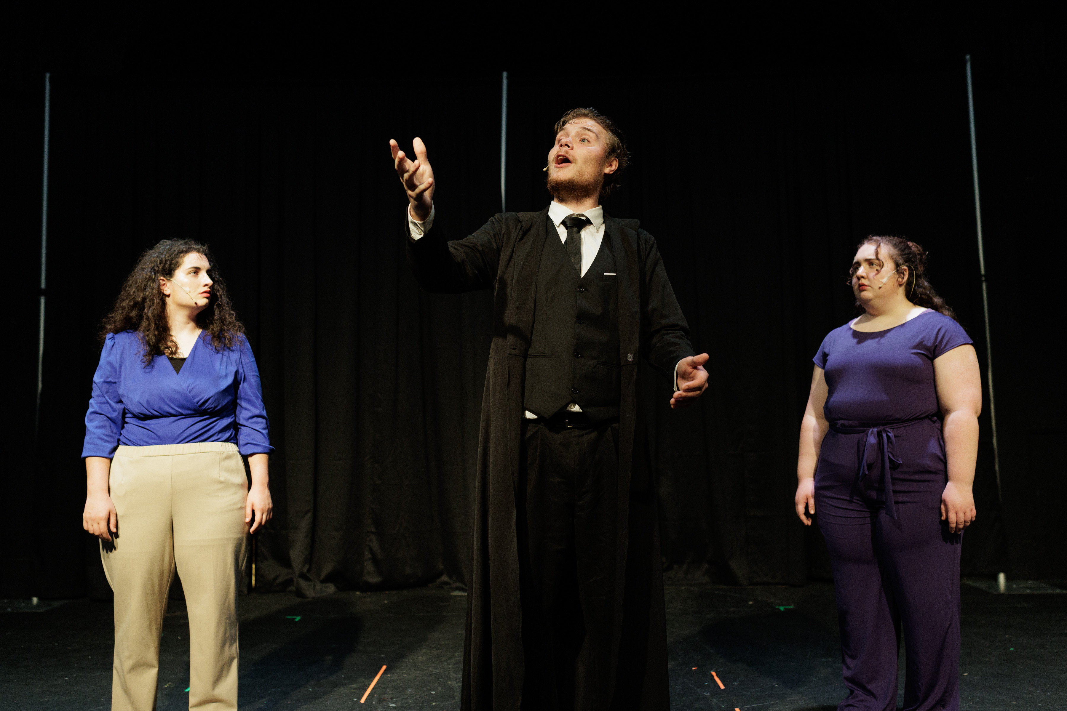 A suited performer raises one hand and looks upward while two performers watch in Two Part Tragedy.
