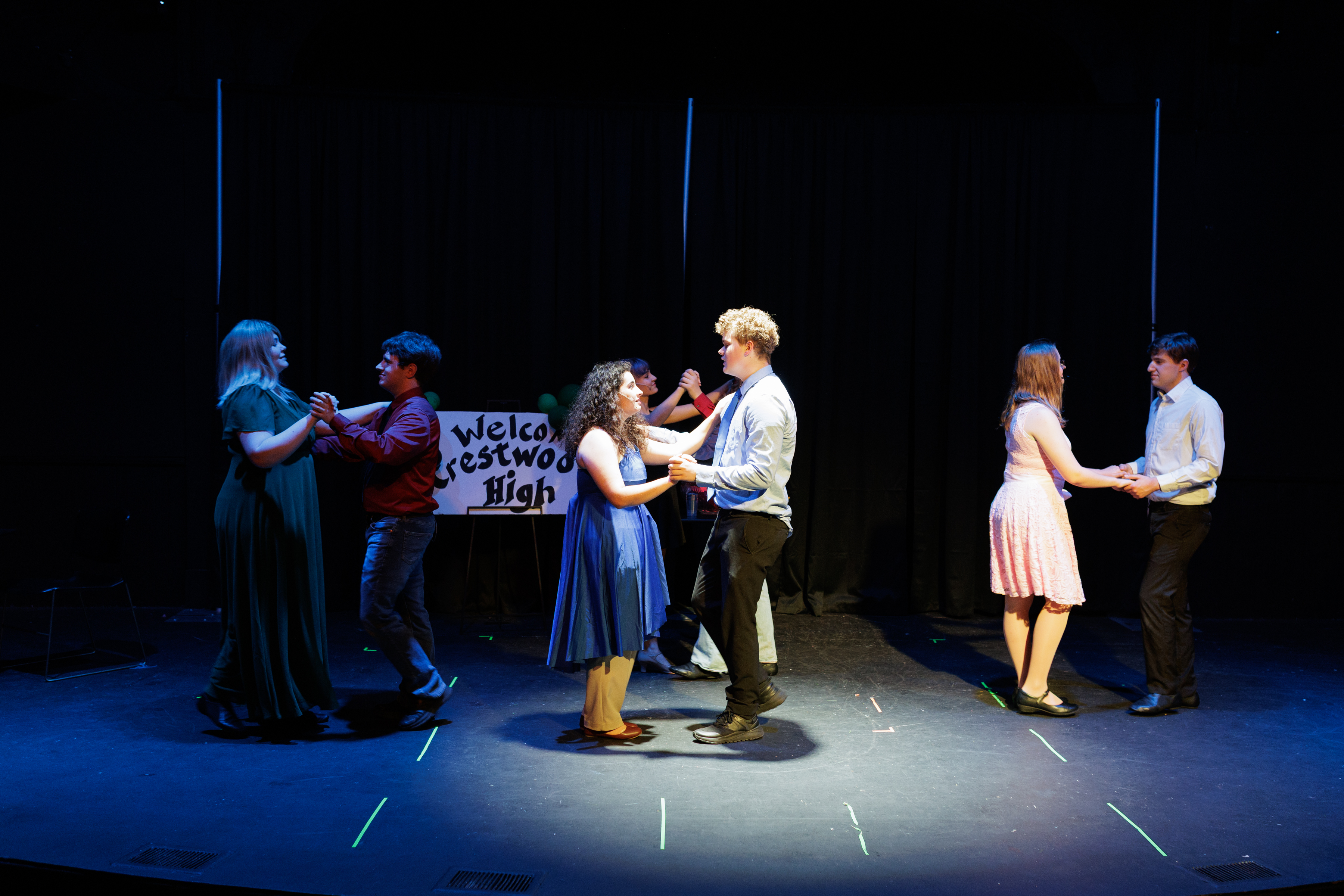 Three pairs dance beneath blue stage lighting beside a school welcome sign in Two Part Tragedy.