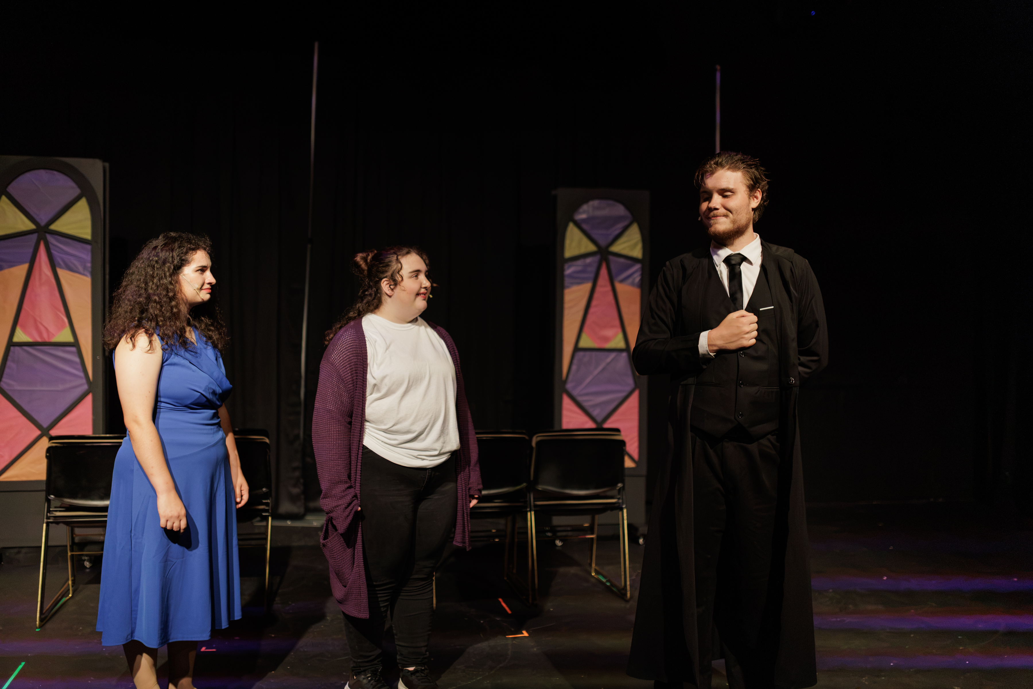 Performers in a blue dress, purple cardigan, and black suit stand before colorful window panels in Two Part Tragedy.