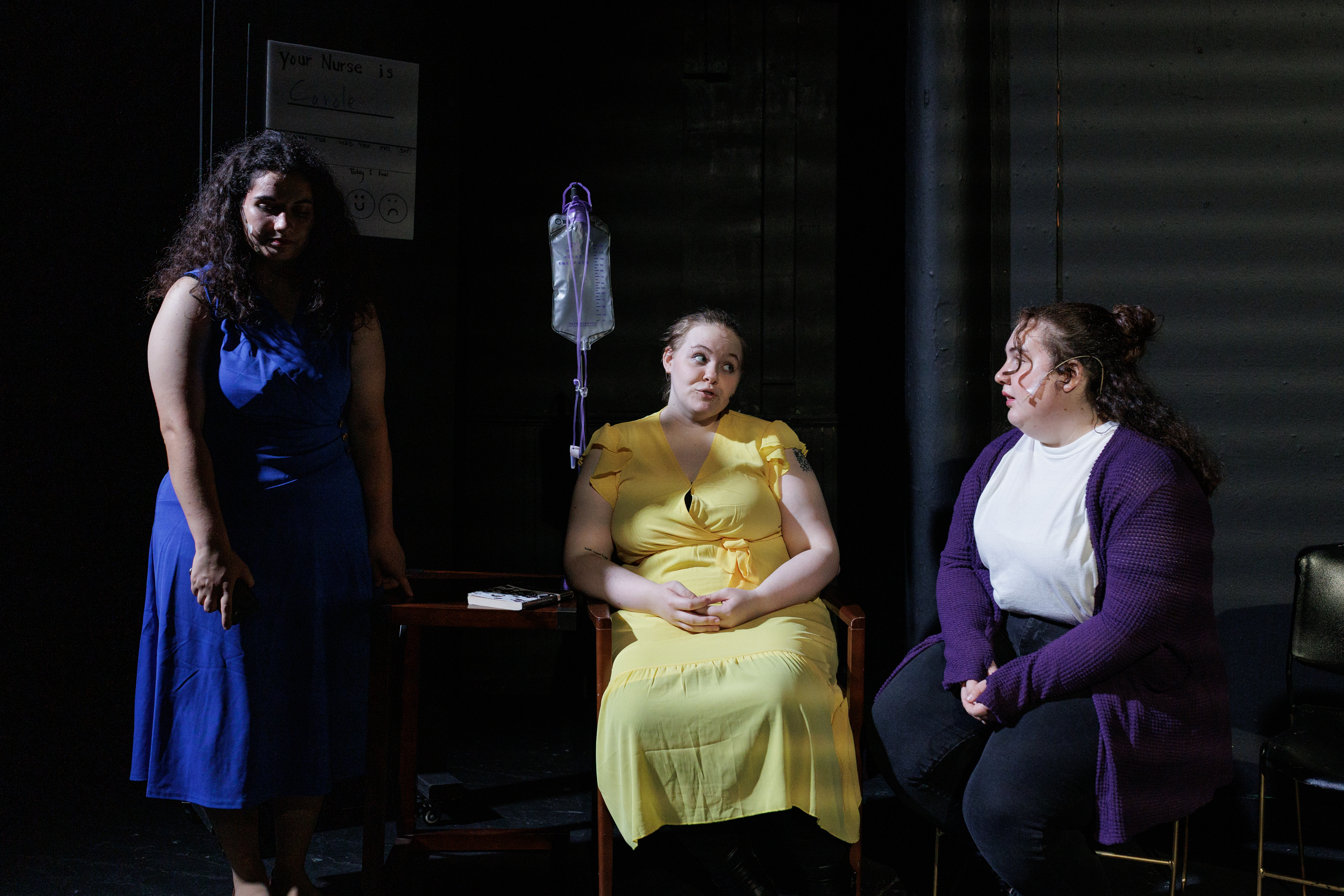 A seated performer in yellow is flanked by performers in blue and purple beside an IV stand in Two Part Tragedy.