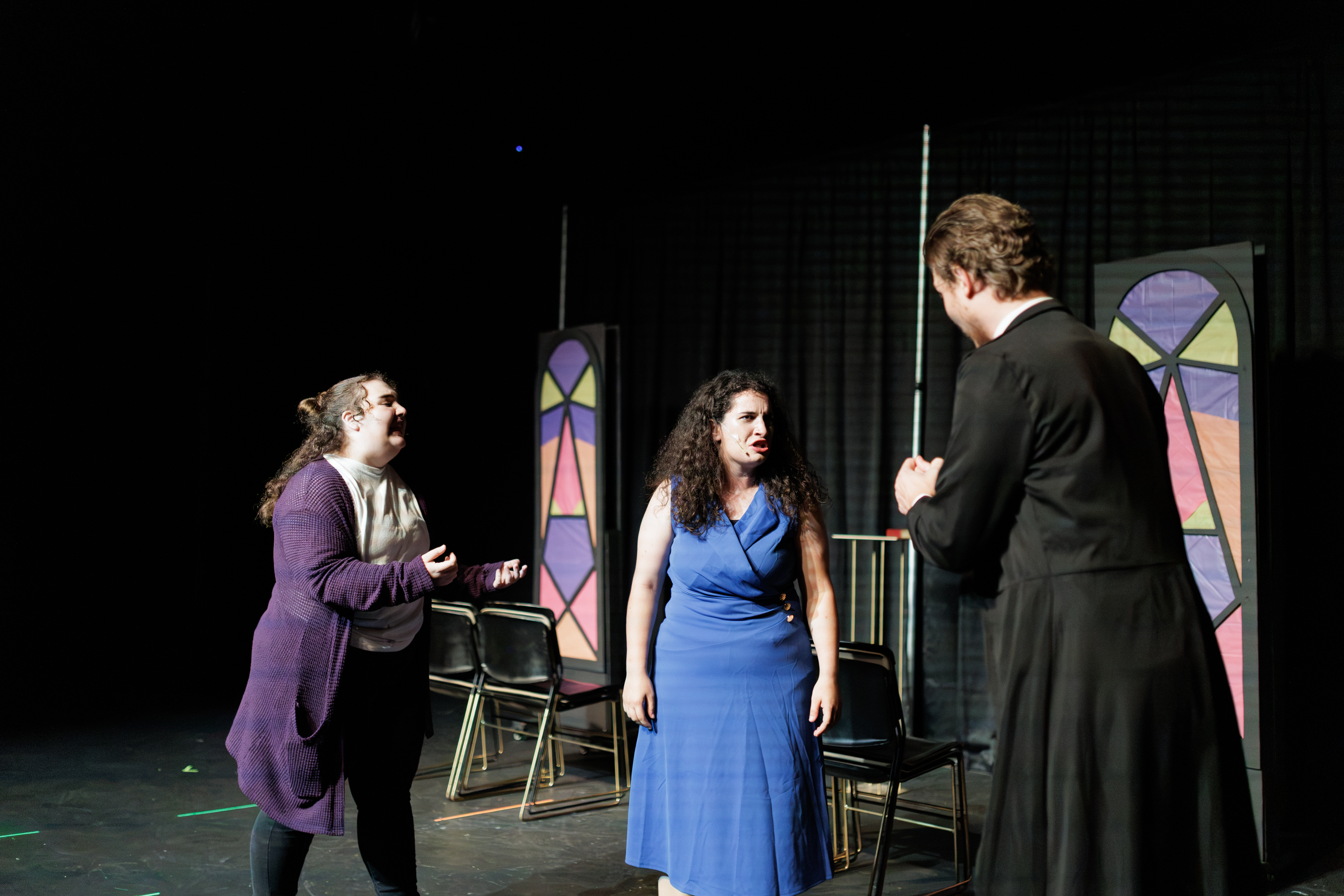 A performer in a purple cardigan gestures with open hands beside performers in blue and black in Two Part Tragedy.