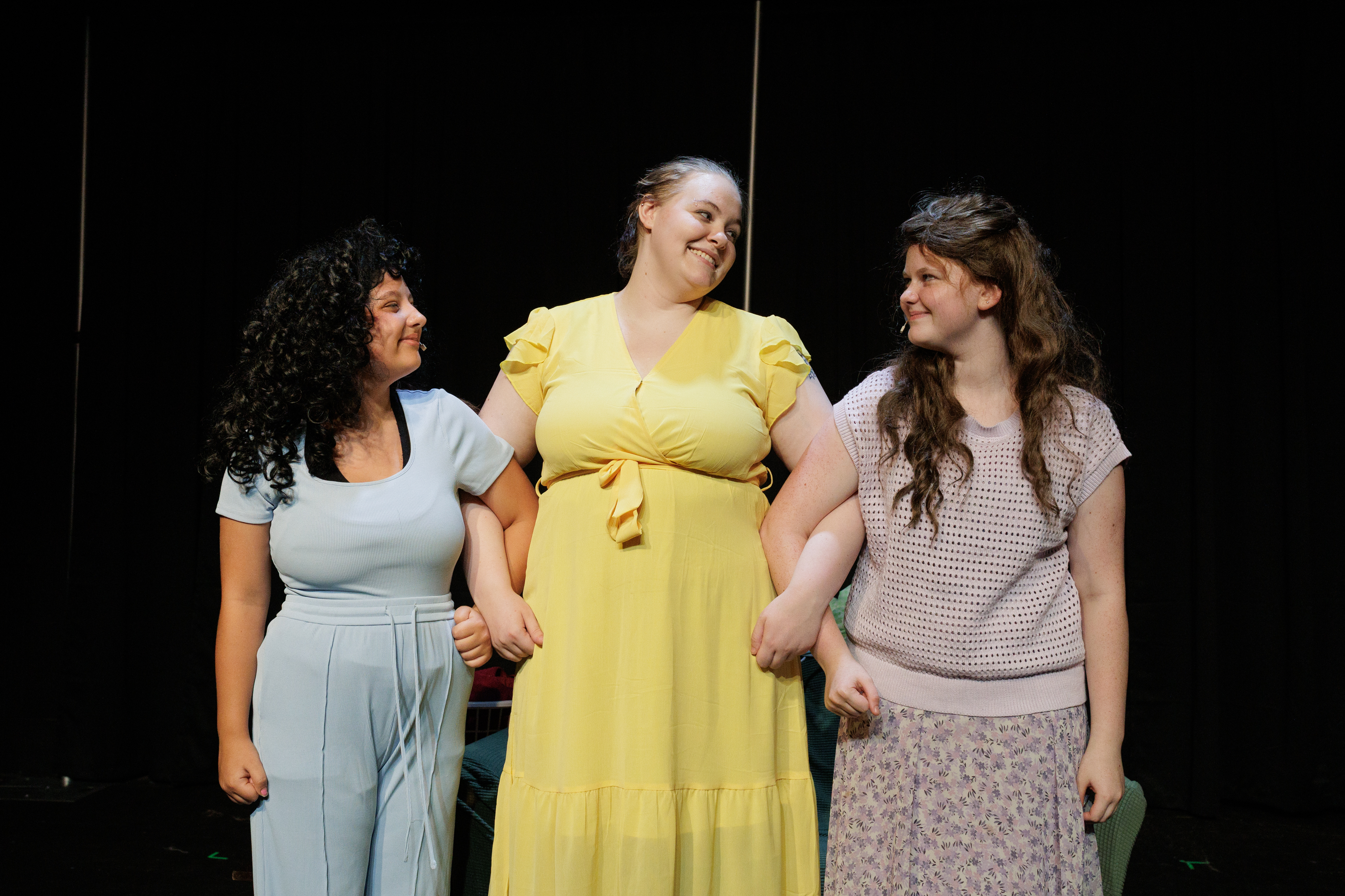 Three smiling performers in light blue, yellow, and lavender link arms in Two Part Tragedy.