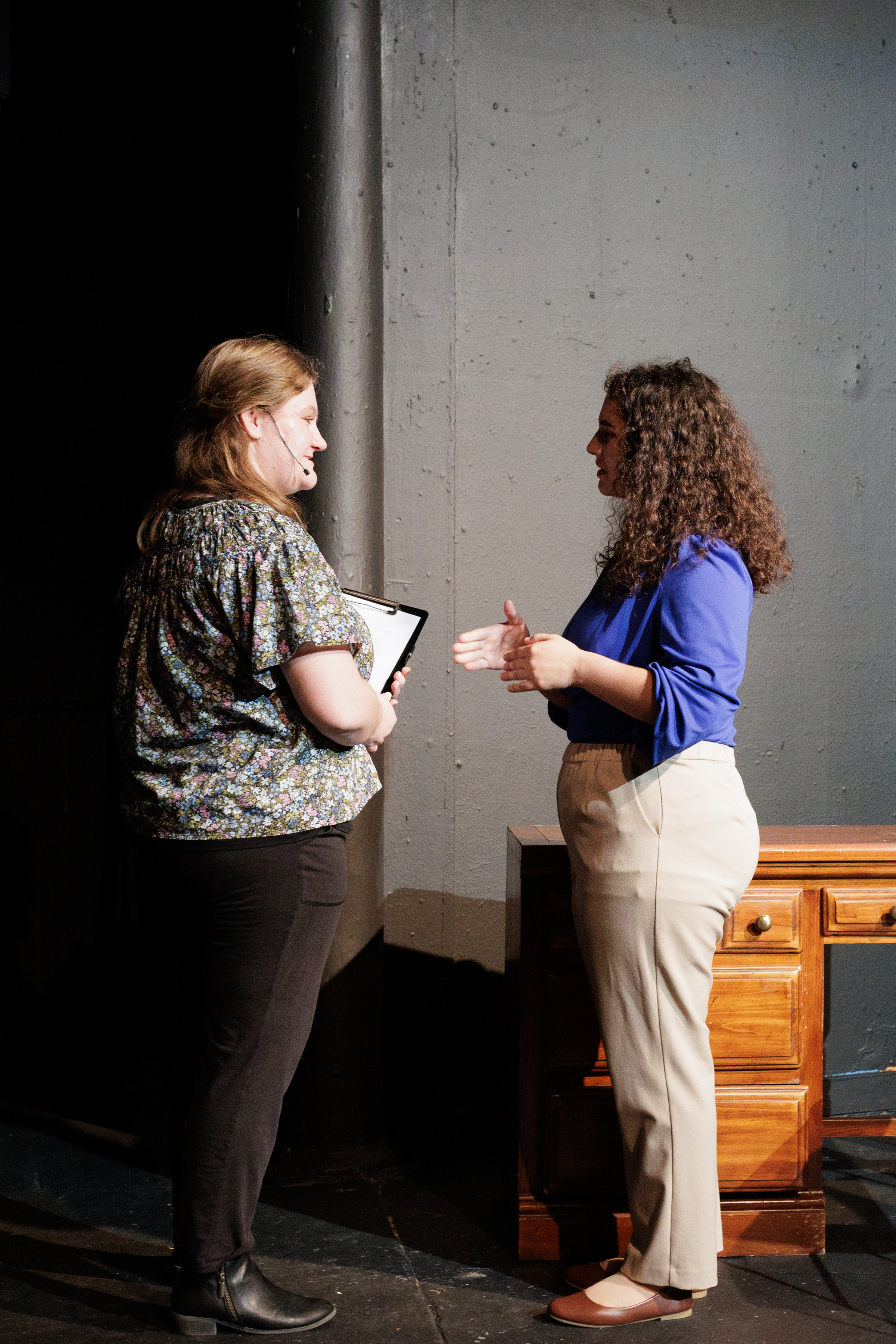 Two performers face each other beside a wooden desk, one holding a folder or clipboard, in Two Part Tragedy.