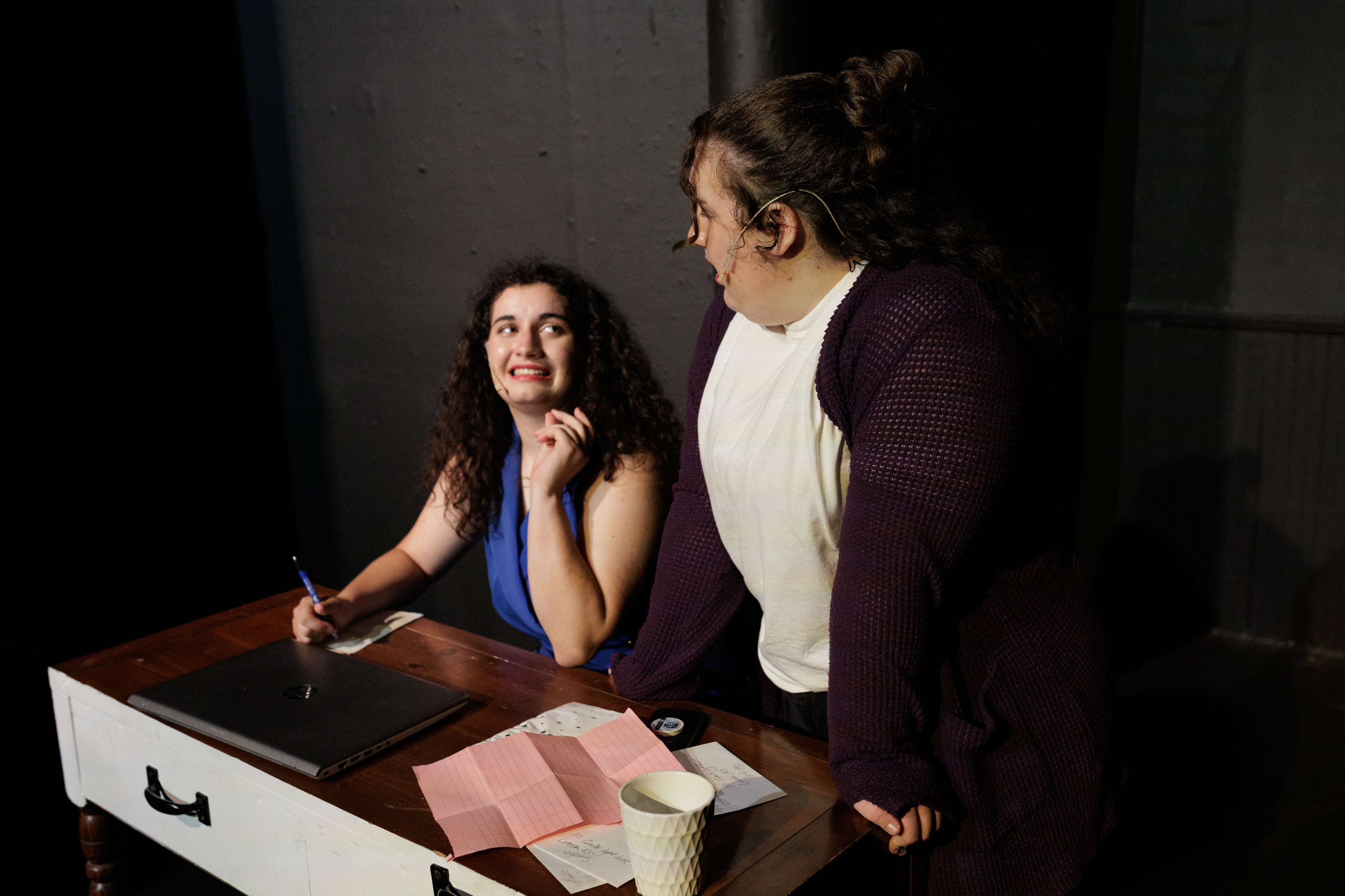 A performer in blue holds a pen over a notepad while looking up at a performer in a purple cardigan in Two Part Tragedy.