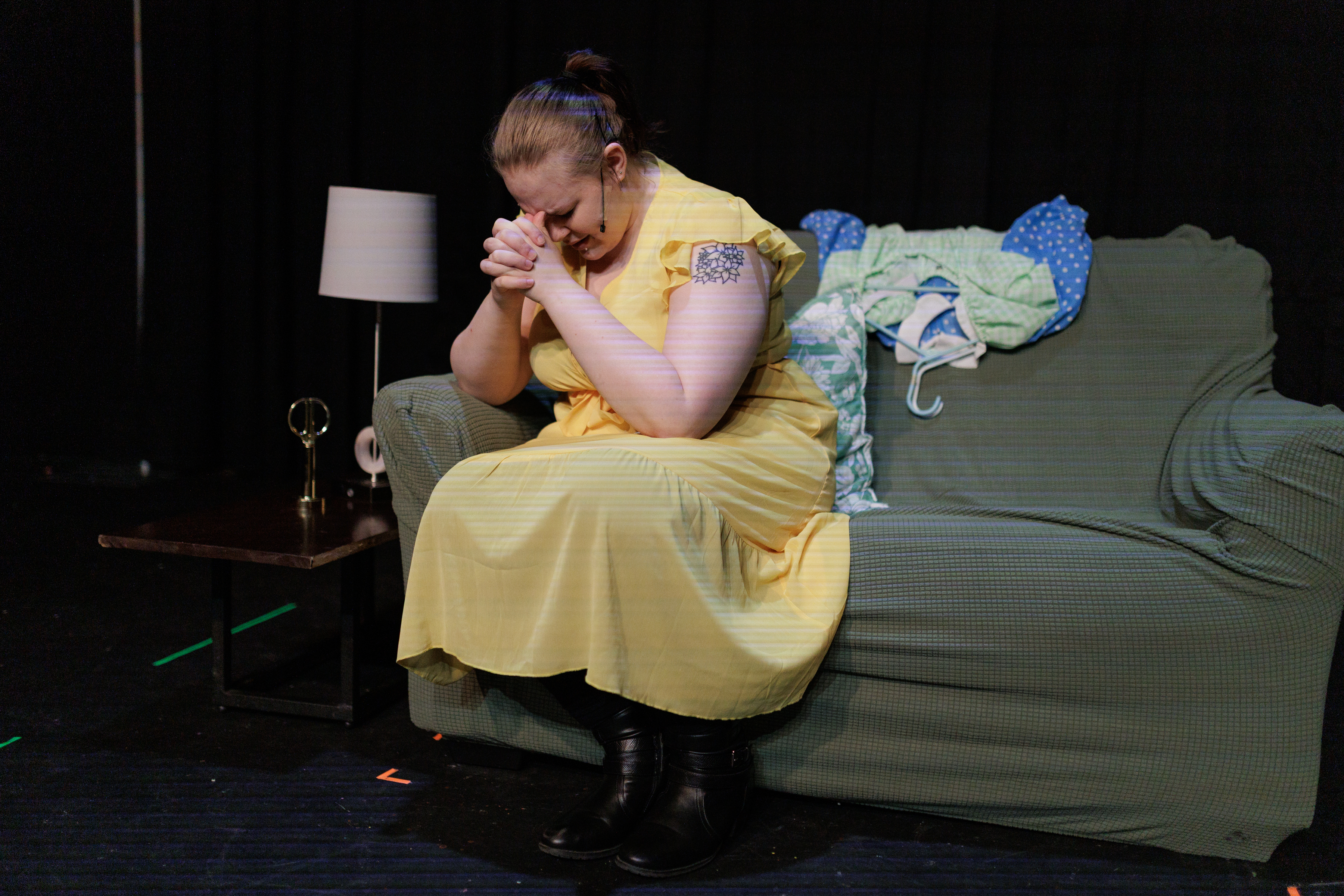 A performer in a yellow dress sits hunched on a green sofa with hands clasped near their face in Two Part Tragedy.