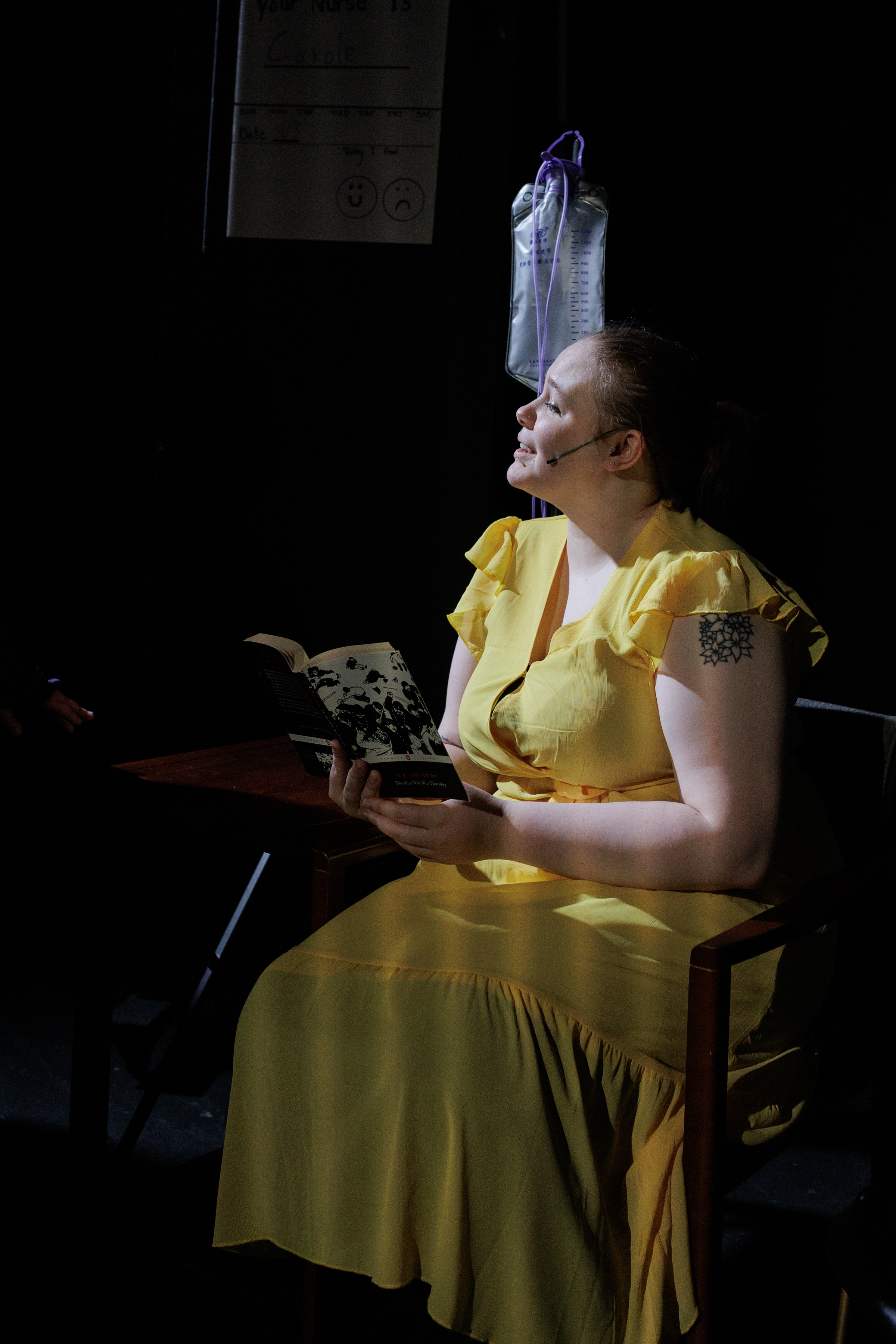 A performer in a yellow dress sits reading a book beside an IV stand in Two Part Tragedy.
